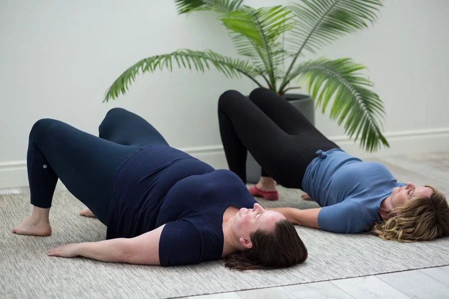 Two women performing a bridge exercise on a yoga mat in a room with a large potted plant.