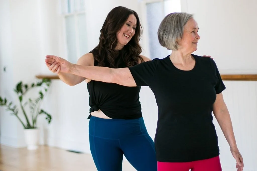 A young woman guides an elderly woman in a stretching exercise in a bright, spacious room with a potted plant in the background.