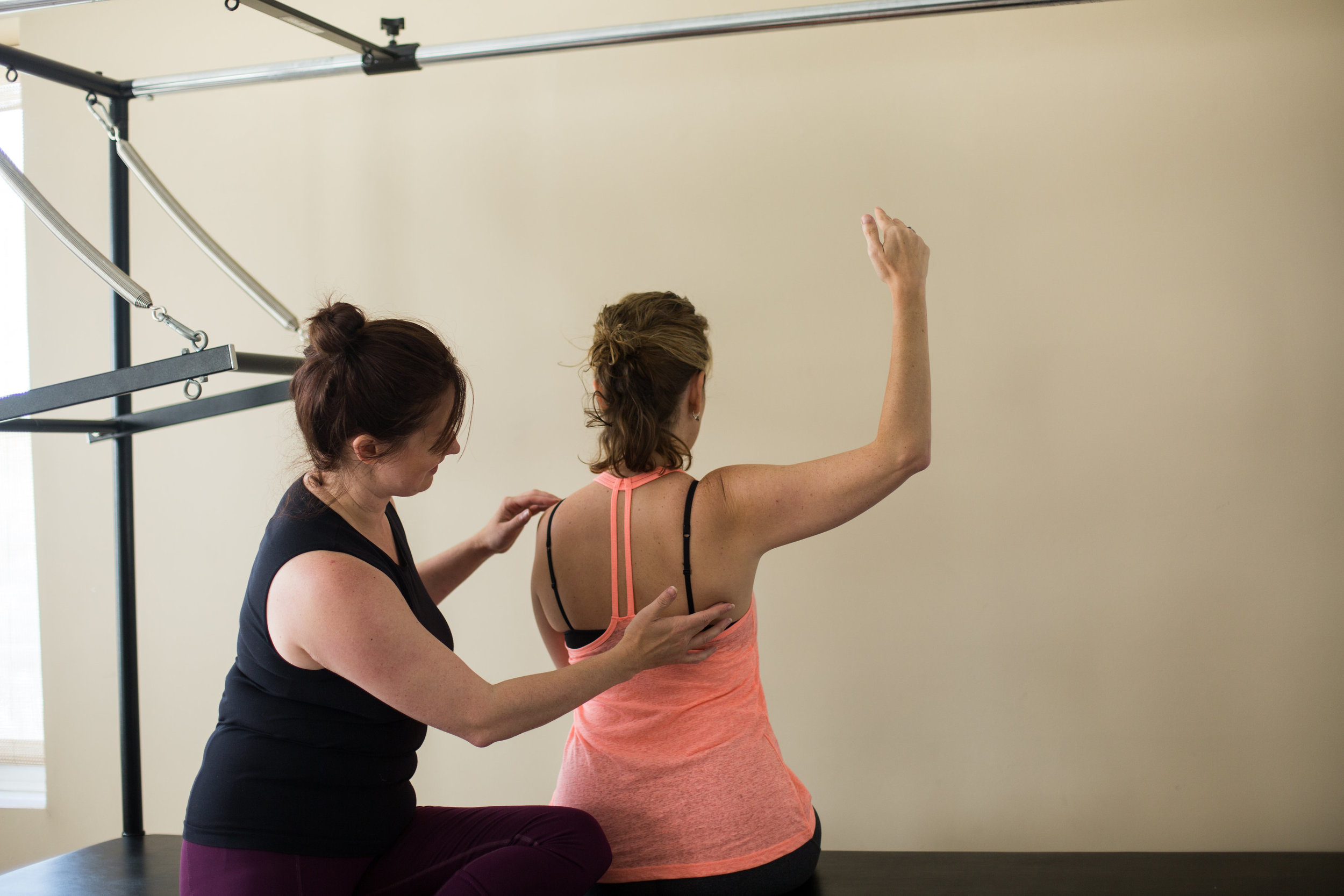 A woman providing physical therapy or injury assessment to another woman, who is standing with her arm raised and back to the camera, in a clinical or gym setting.