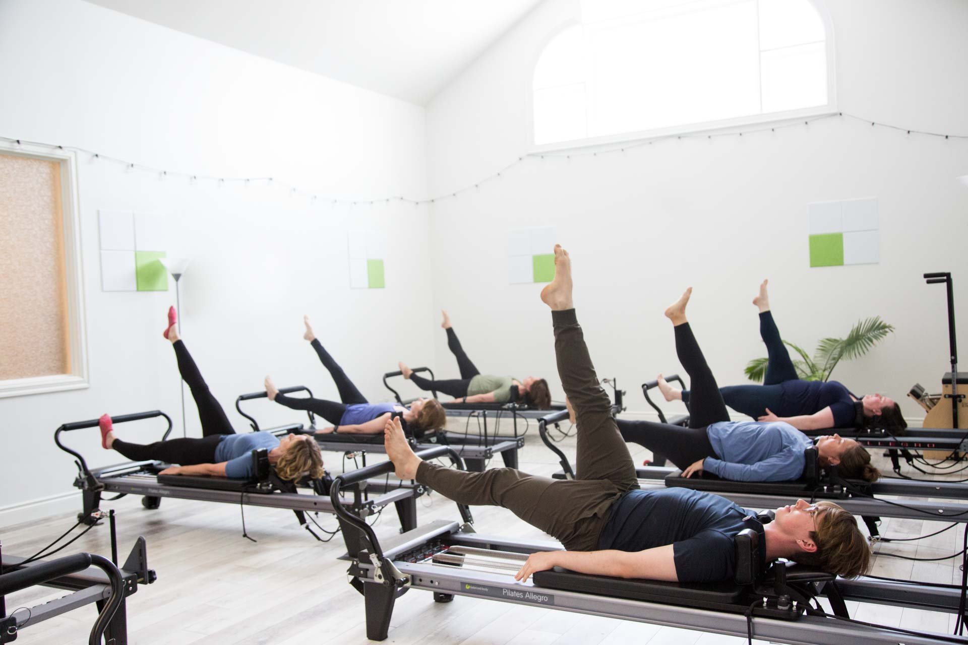 Group of people exercising on Pilates reformer machines in a bright studio with white walls and high windows.