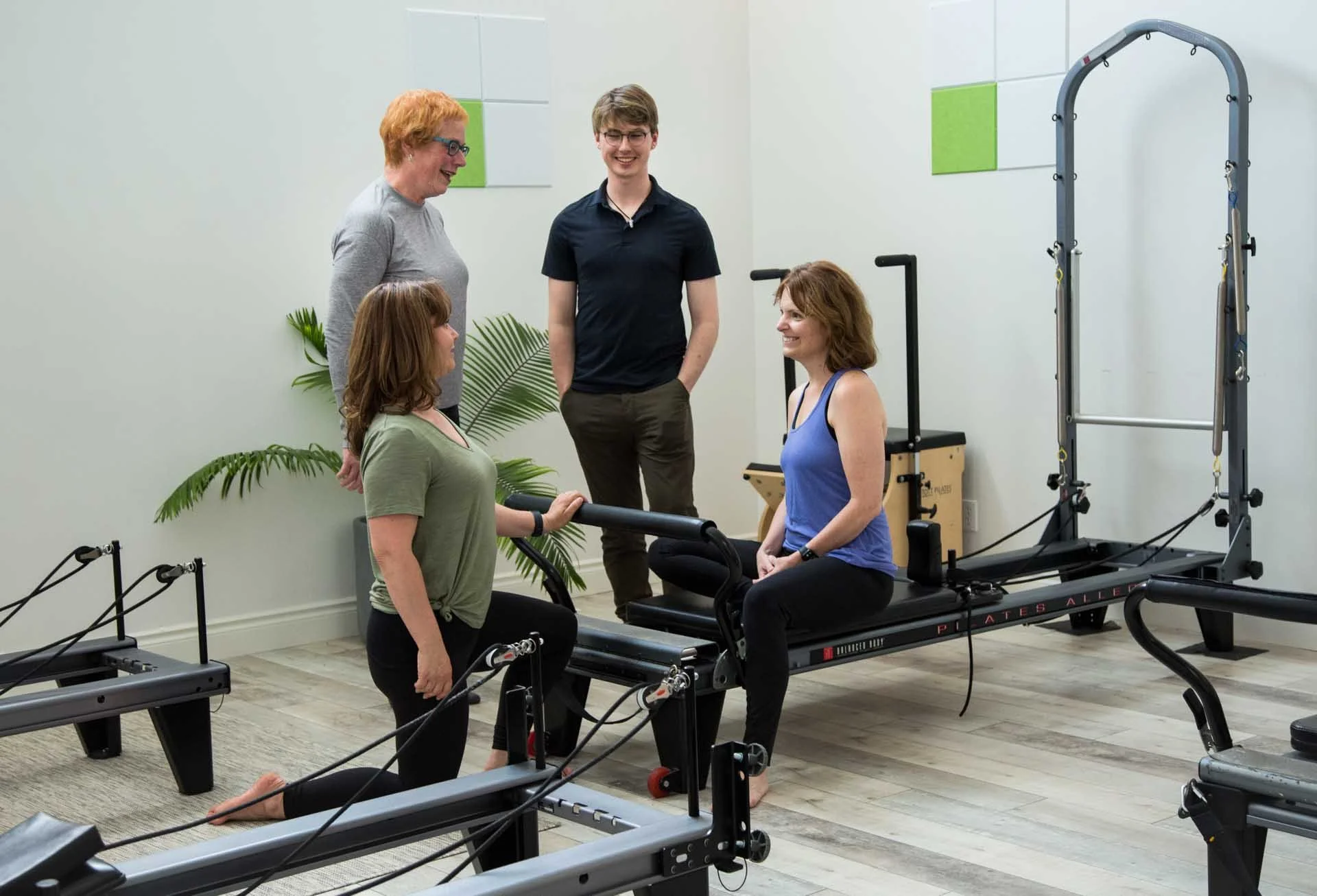 A group of five people in a fitness or physical therapy room with Pilates reformer machines. Two women are seated on reformers, one standing next to her, and one is crouching next to a reformer. A man and a woman are standing, smiling and talking, while another woman looks on. There are green and white wall panels and some plants in the background.