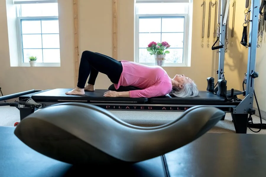 An elderly woman doing a bridge exercise on a Pilates reformer machine in a well-lit room with windows, a potted plant, and workout equipment.