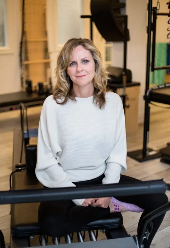 A woman with blonde hair and a white sweater sitting on a black Pilates reformer machine in a fitness studio with equipment in the background.
