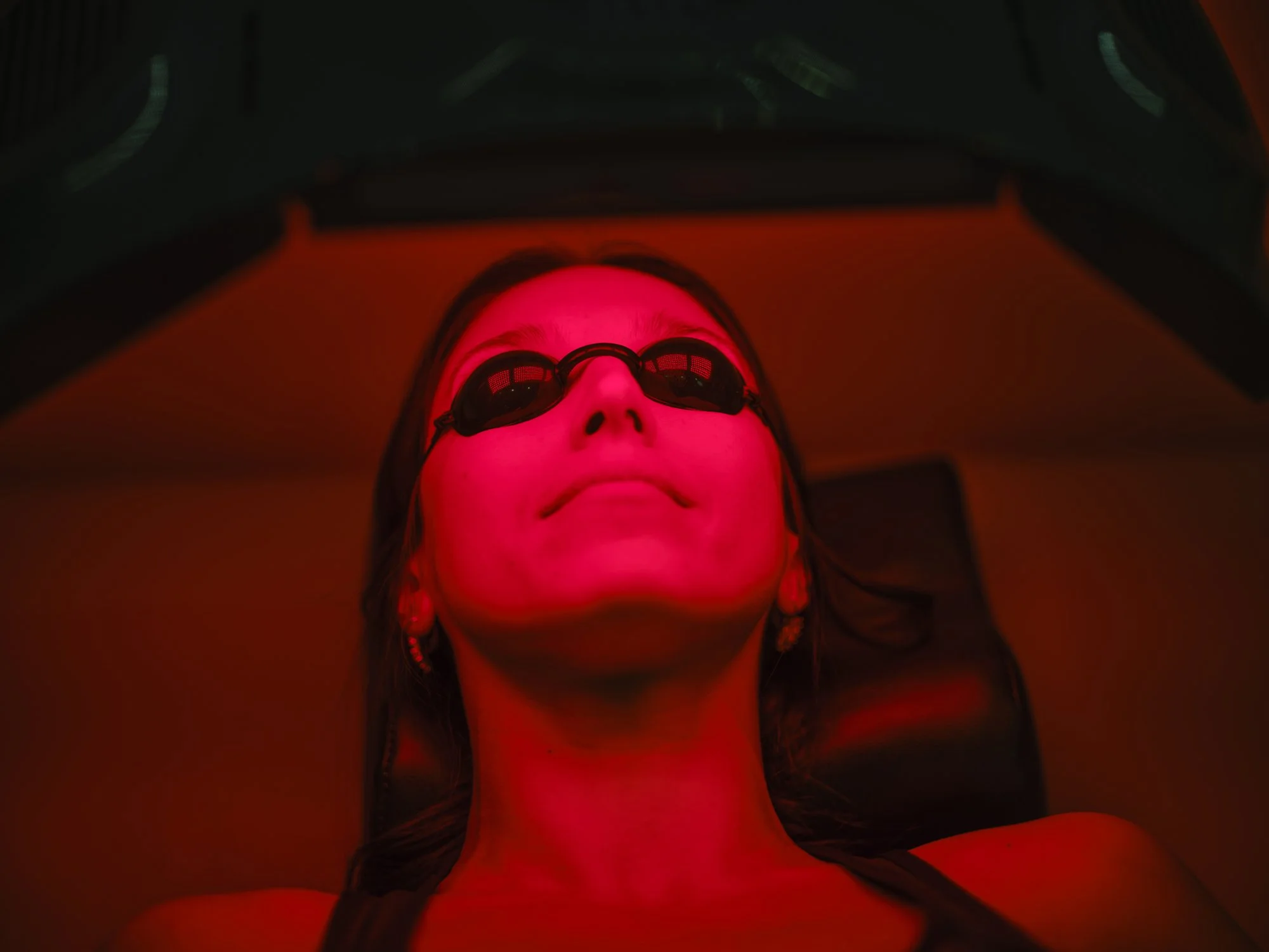 A woman appears to be getting a red light therapy treatment, lying on a surface with her face illuminated by red light and wearing glasses.