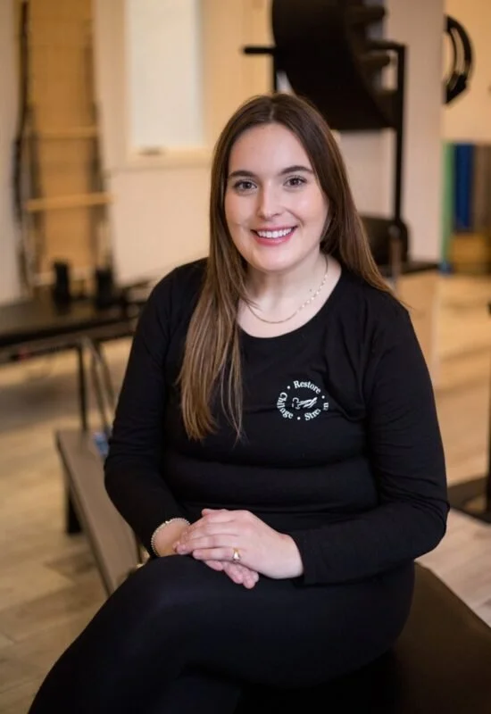 A smiling young woman with long brown hair sitting on a massage table in a wellness or physical therapy center.
