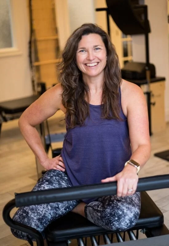 A woman with long brown curly hair sitting on a black exercise bench in a fitness studio, smiling at the camera, wearing a purple tank top and patterned leggings.