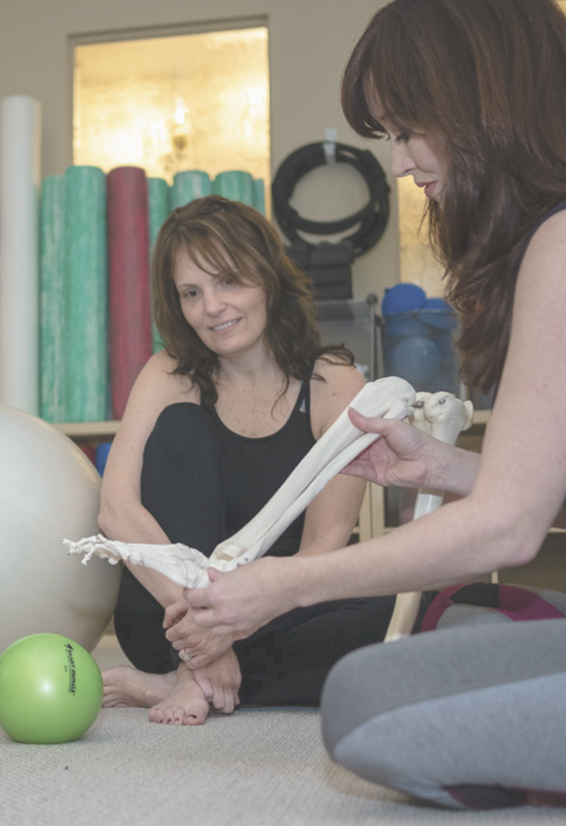 Two women sitting on the floor in a fitness or therapy room, one is holding a large anatomical model of a leg bones, the other is watching. There are yoga and exercise balls and fitness equipment in the background.