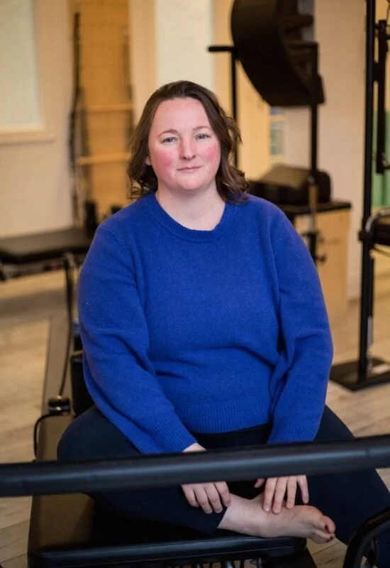 A woman with shoulder-length brown hair, wearing a blue sweater, sitting on a black chair in a room with calm lighting.