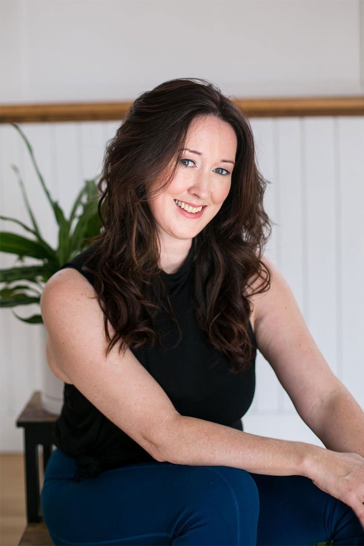 A woman with long, dark, wavy hair smiling, sitting in front of a white wall with a wooden ledge and a green plant in the background.