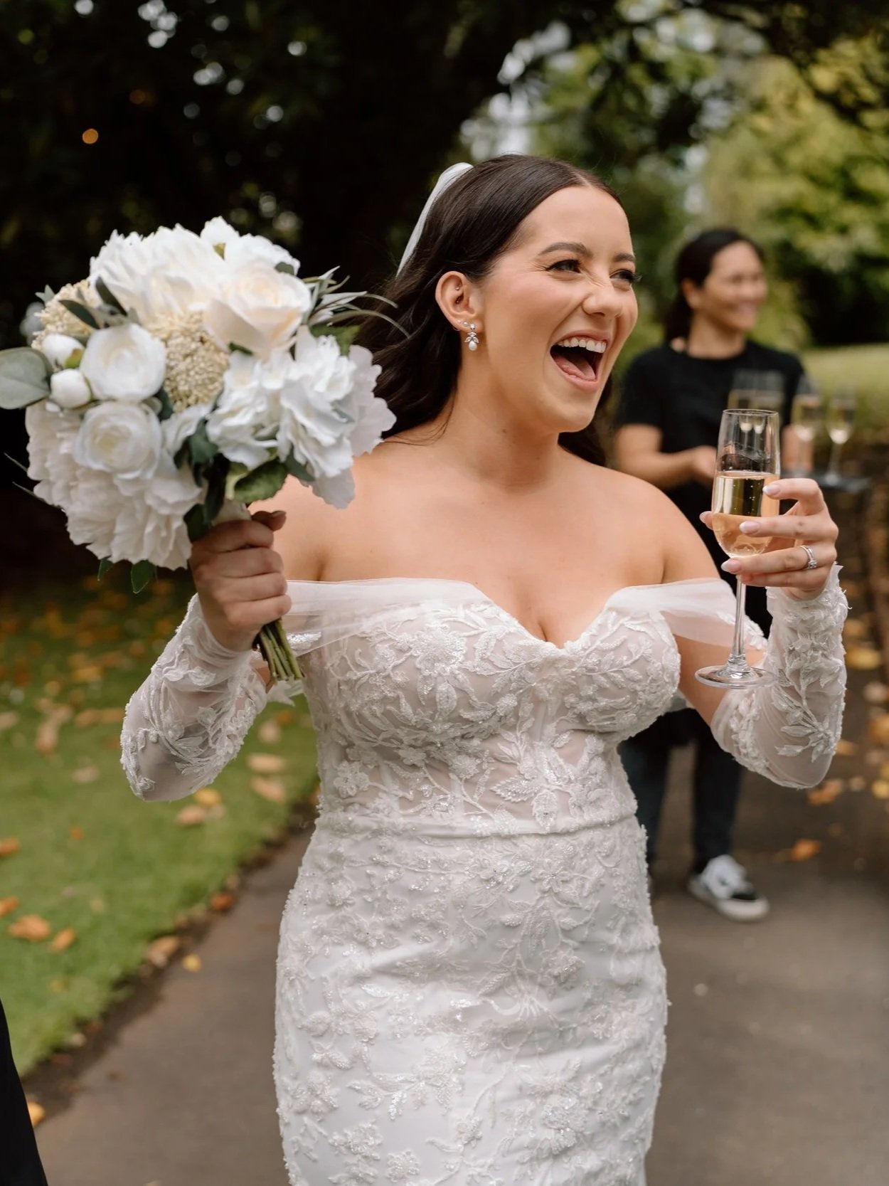 Joyful bride in a lace wedding dress holding a bouquet of white flowers and a glass of champagne during outdoor celebration with guests.
