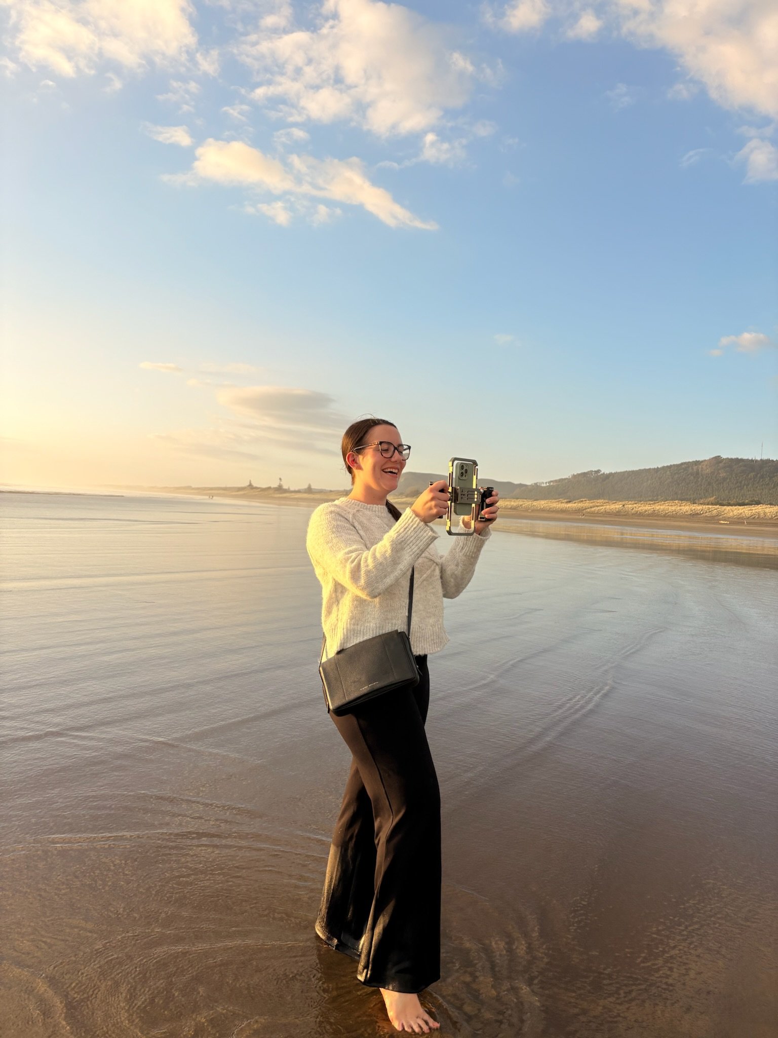 A woman with glasses standing in shallow water at the beach, smiling and taking a photo with her phone during sunset.
