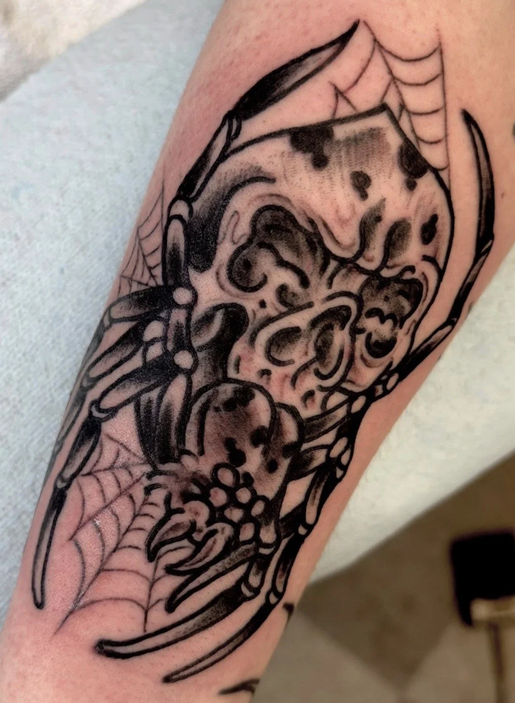 Close-up of a black-and-gray tattoo featuring a stylized skull, flowers, spiderwebs, and decorative scrollwork on an arm.