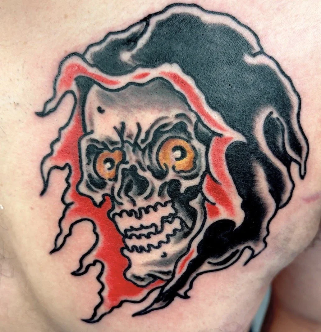 Close-up tattoo of a skull with orange eyes beneath a dark hood, surrounded by red flames.