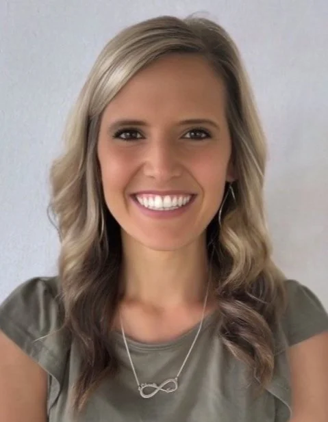 A woman with long, wavy blonde hair, smiling, wearing a gray top and a silver infinity necklace.