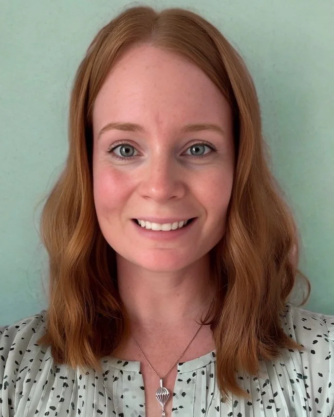 A woman with red hair and blue eyes smiling in front of a light green background, wearing a white blouse with black polka dots and a necklace.