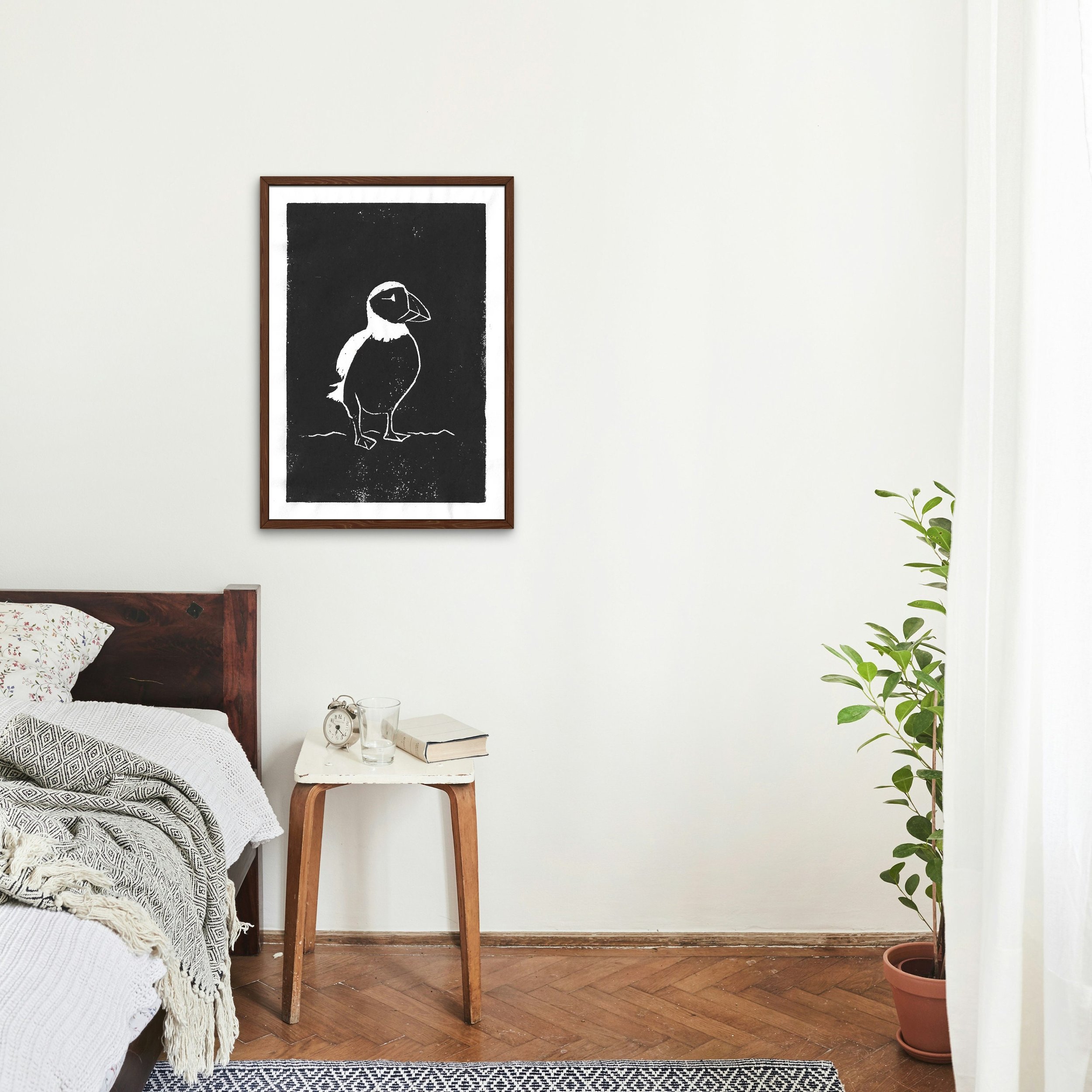 A puffin lino cut print on a white wall, styled above a bedside table and soft, neutral bedding.