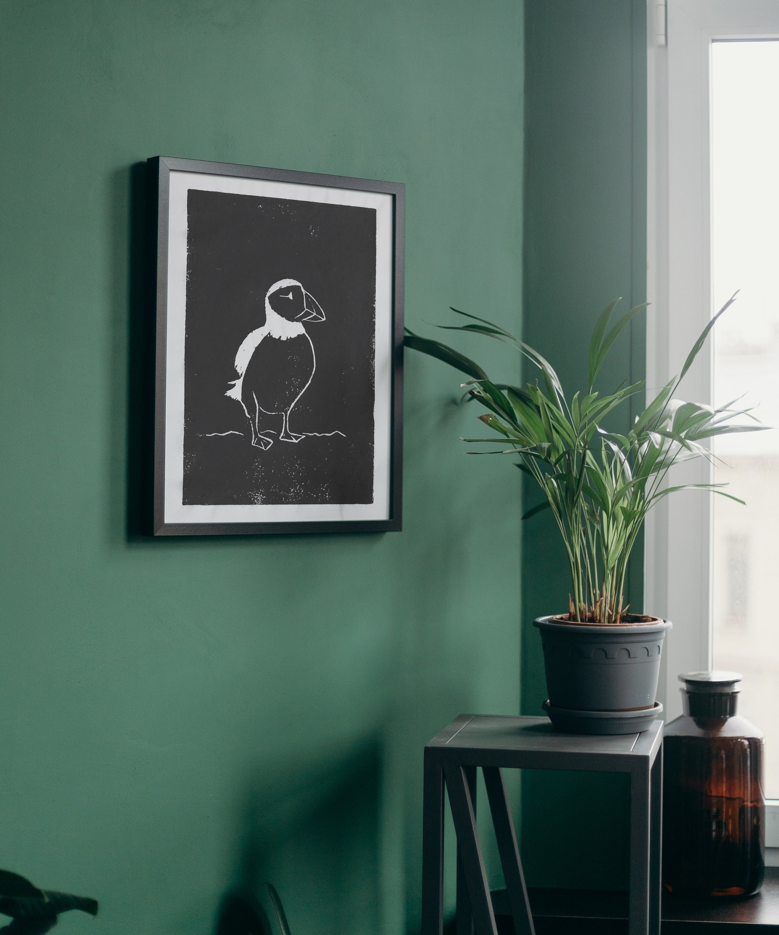 A framed puffin print displayed on a bold green wall with a minimalist shelf and plants.