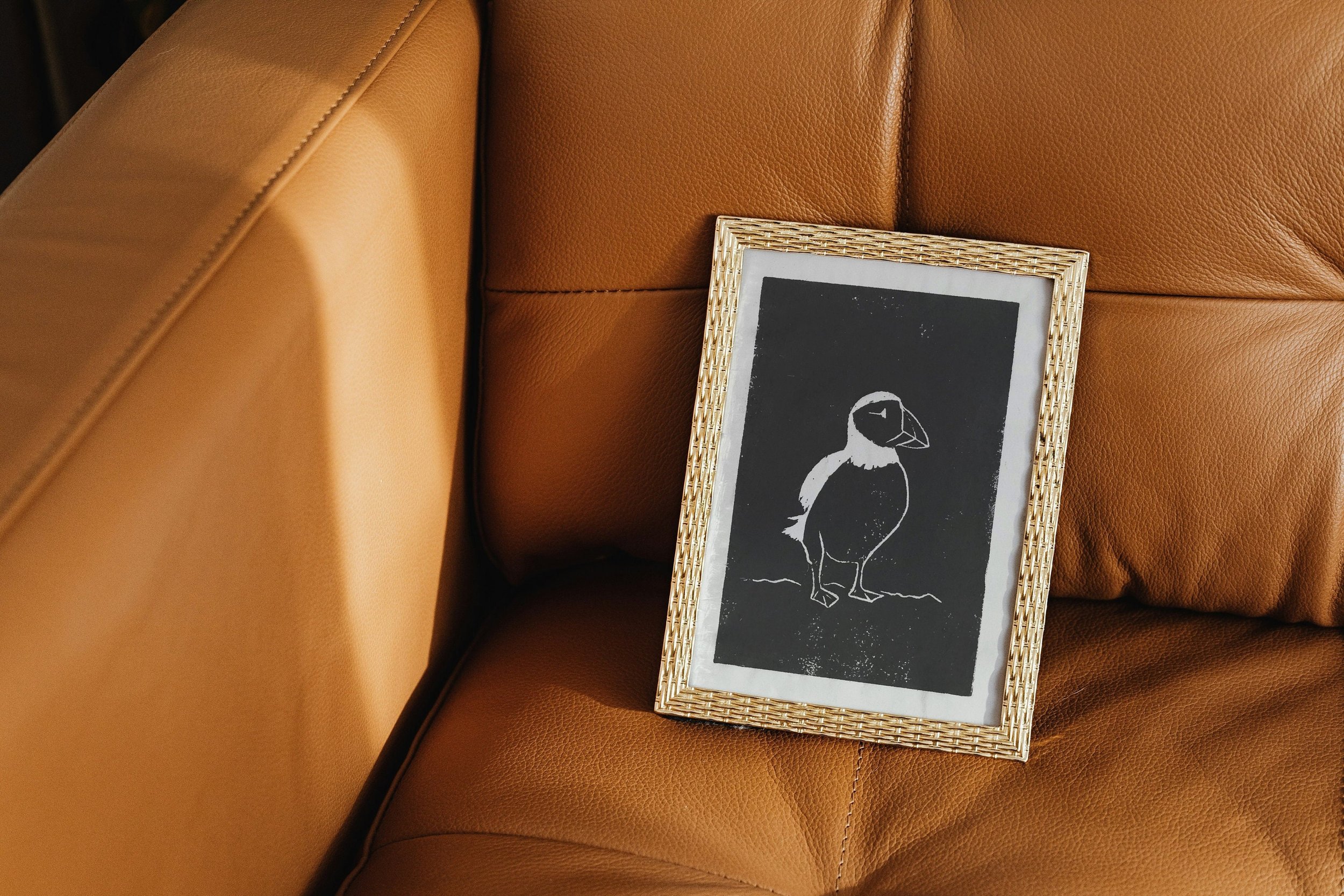 A puffin lino cut print in a gold frame resting on caramel-toned leather furniture.