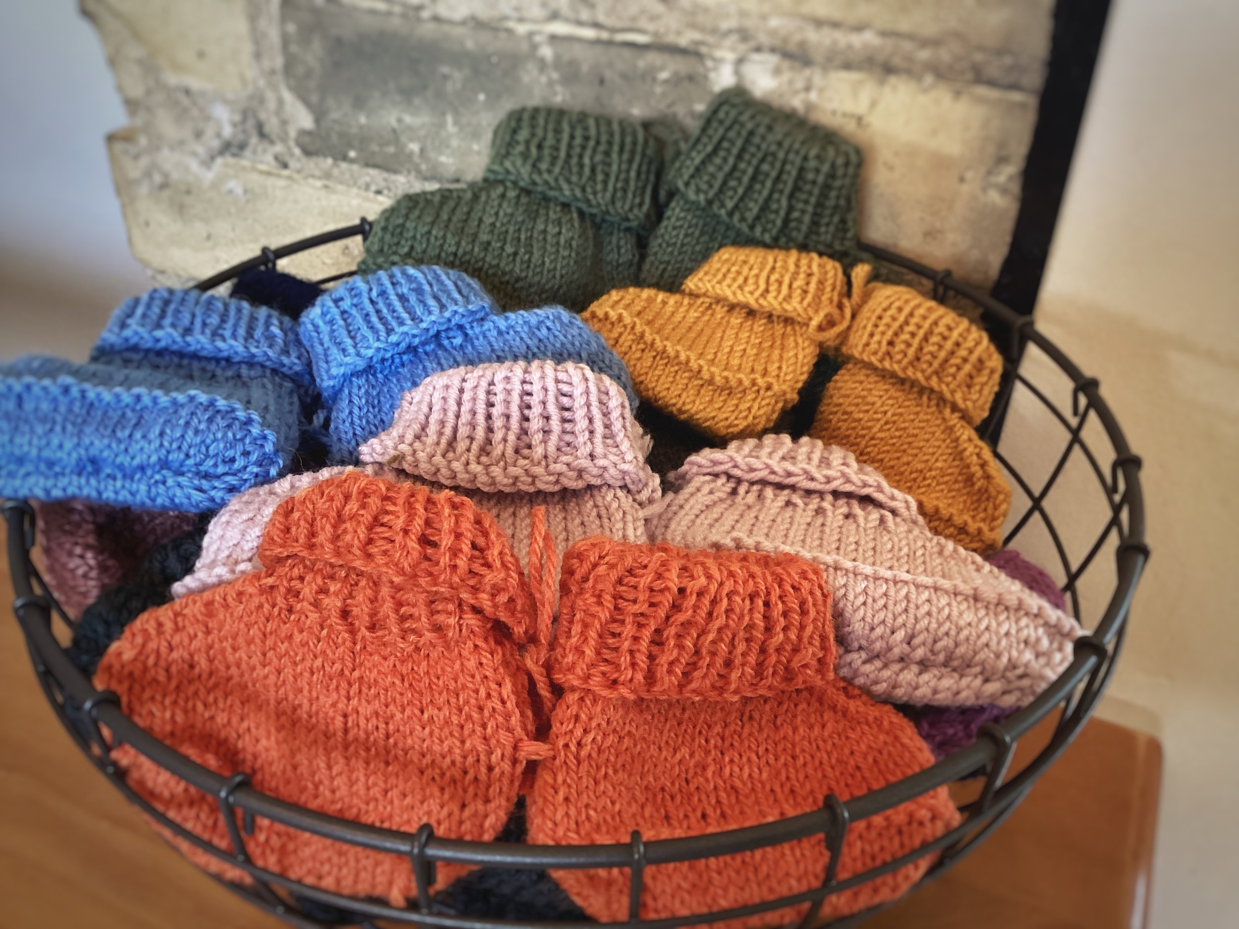 A basket of cozy, handcrafted baby booty slippers in multiple colors, made with a soft wool blend. Ideal for keeping little feet warm.