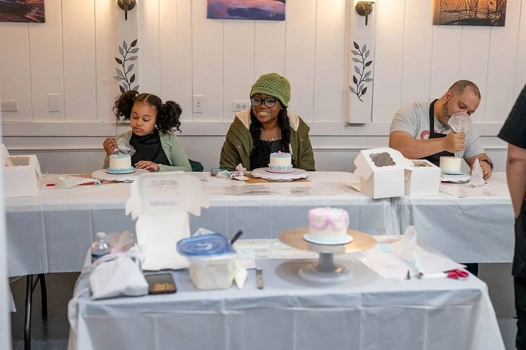 Guests decorating cakes at tables with white linens during a cake class, framed art on the paneled walls behind