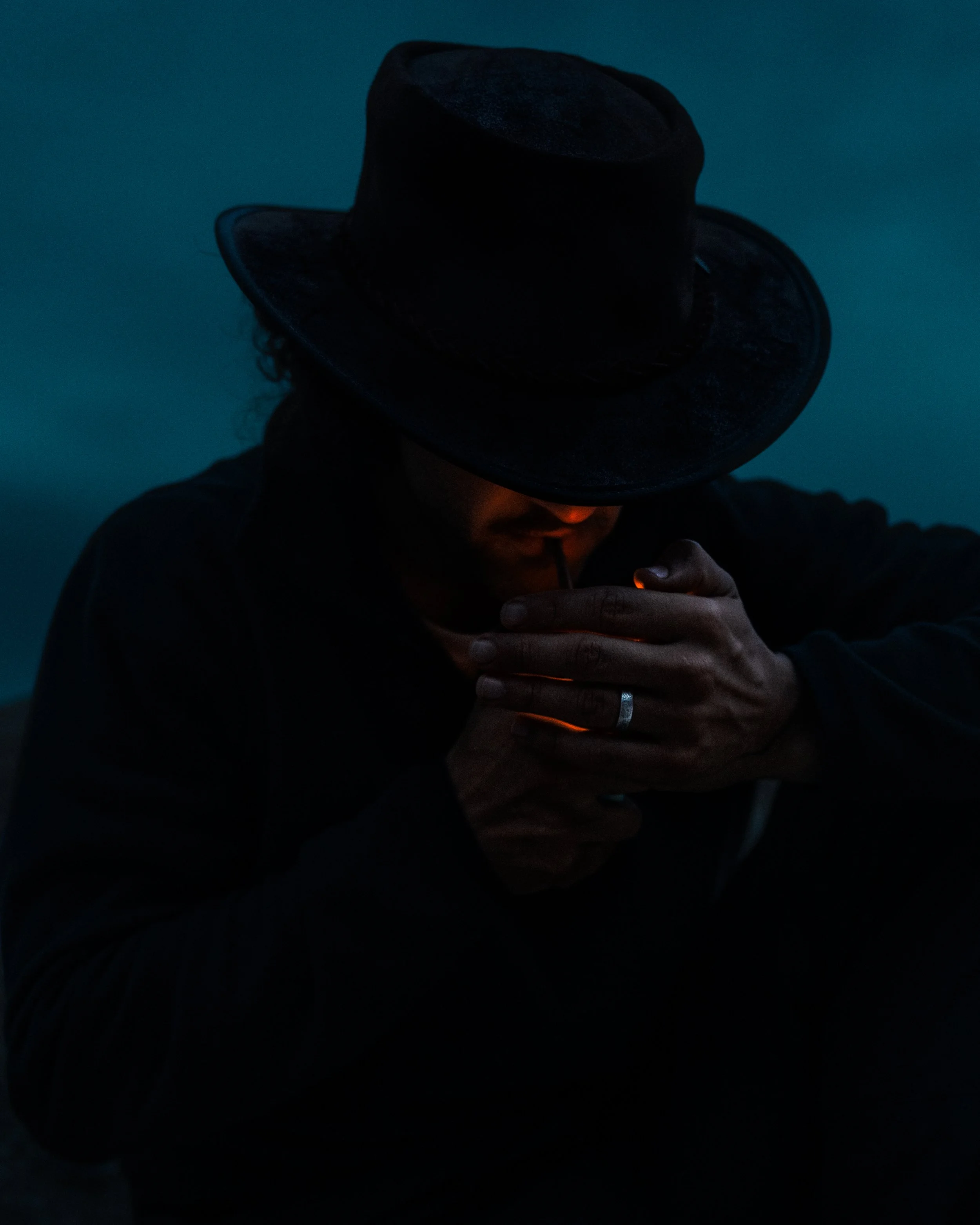 Person wearing a wide-brimmed cowboy hat and dark clothing, holding a lit lighter in front of their chest in a dark setting.