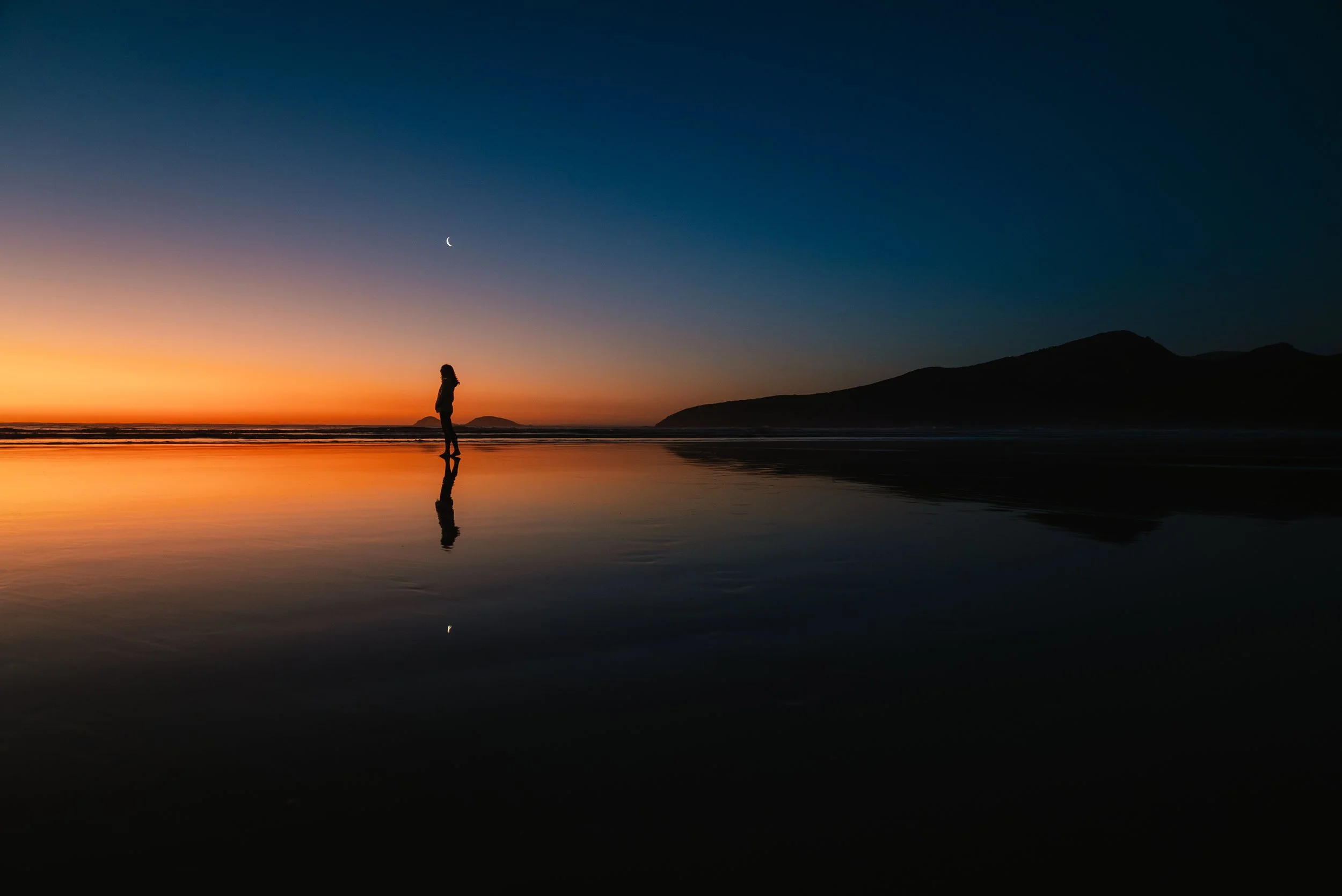 A person standing on a beach during sunset, with a crescent moon visible in the sky and mountains in the background, their reflection visible on the wet sand.