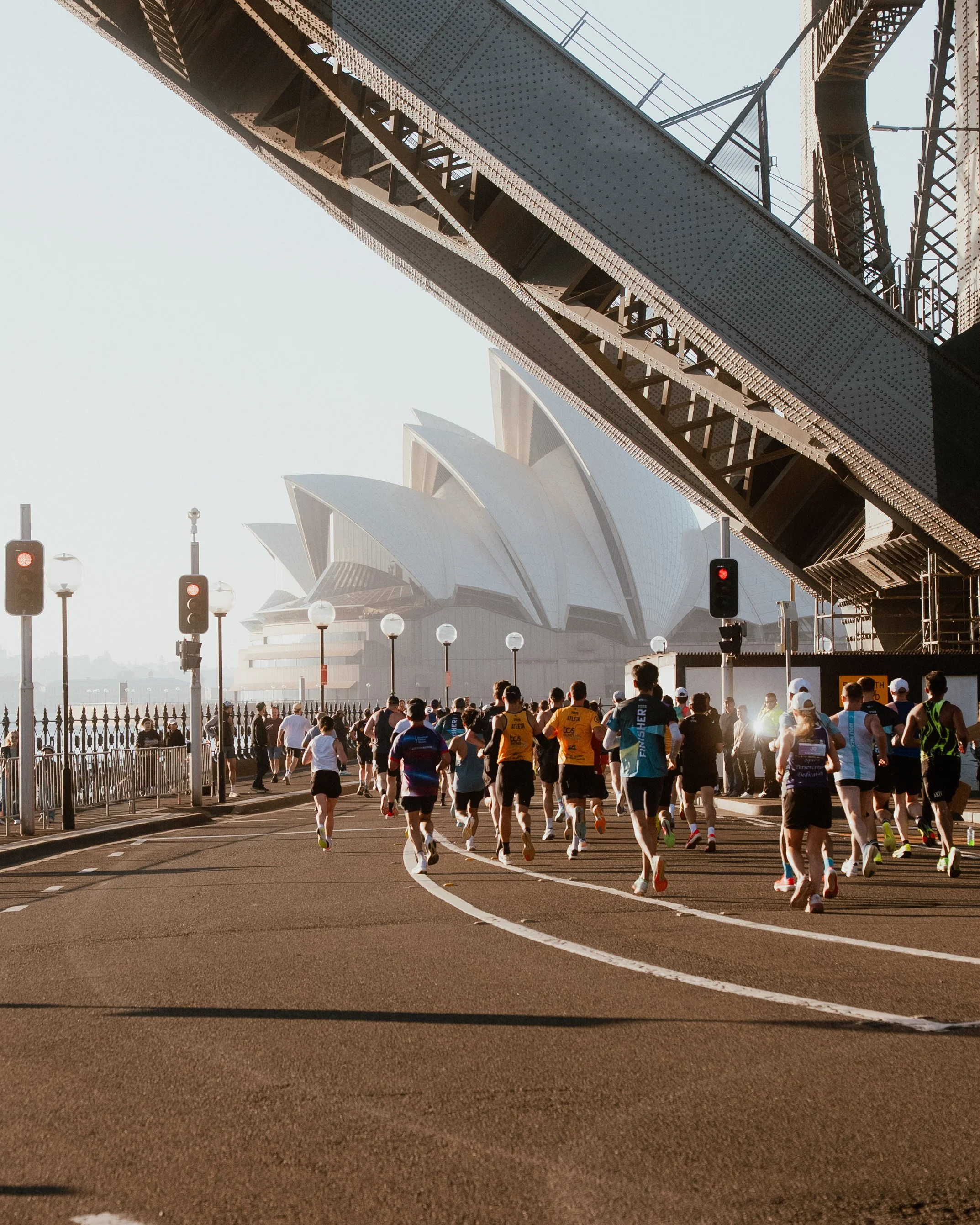 Group of marathon runners running on city street near Sydney Opera House and Harbour Bridge, with traffic lights and street lamps.