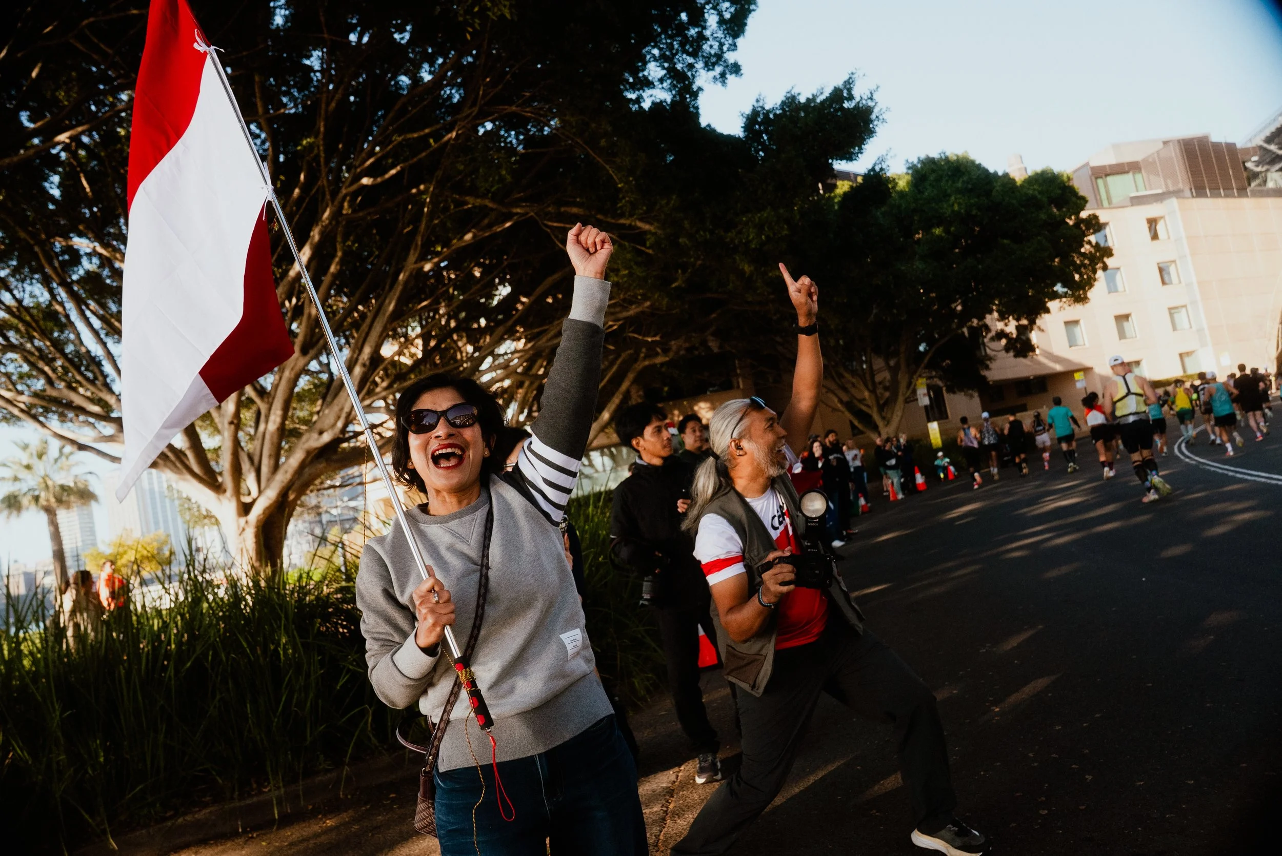 A woman holding a flag with one arm raised, wearing sunglasses, surrounded by a group of people in an outdoor setting with trees and buildings in the background.