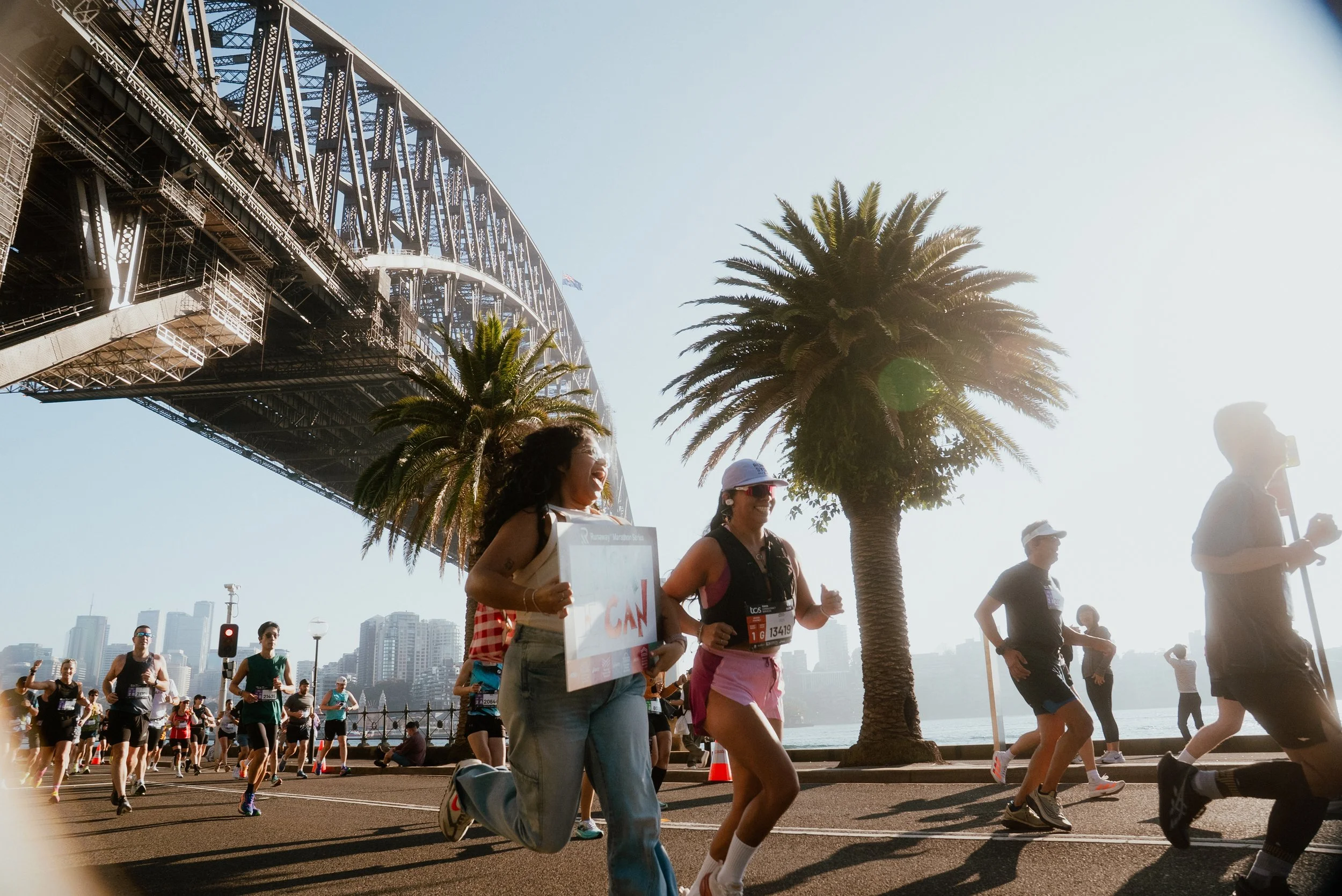 People participating in a marathon race along a waterfront promenade with a bridge and city skyline in the background. A woman in the foreground is smiling and holding a sign. There are palm trees and spectators along the route.