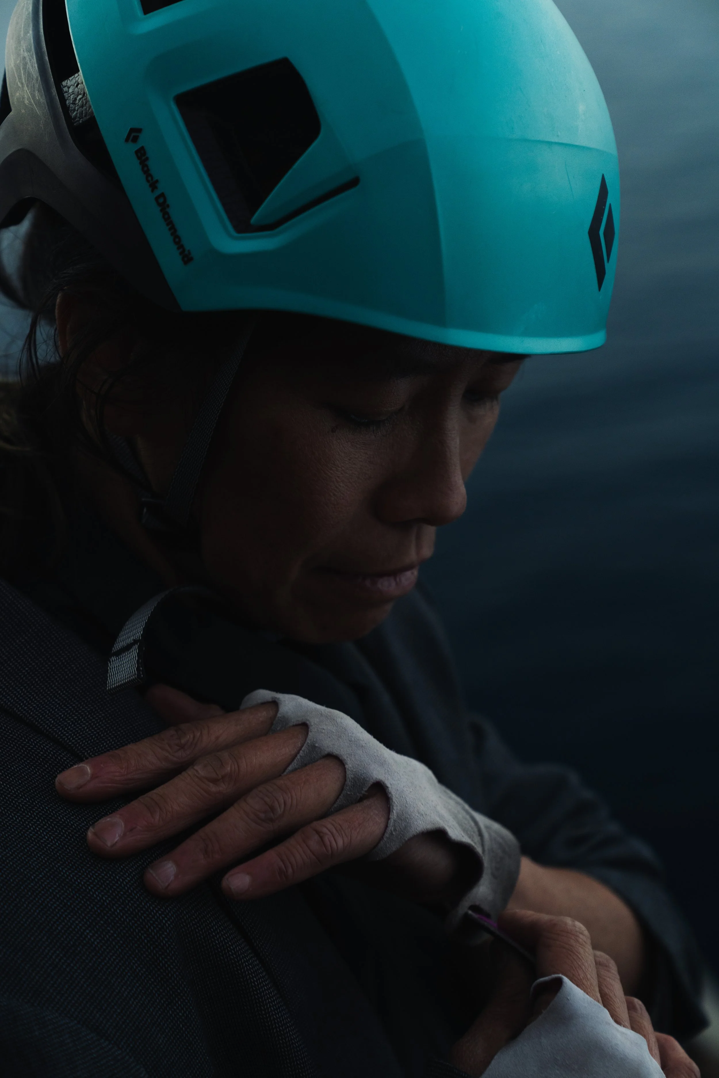 Close-up of a person wearing a blue helmet with the Black Diamond logo, resting their head and hand on another person's shoulder against a blurred blue-gray background.