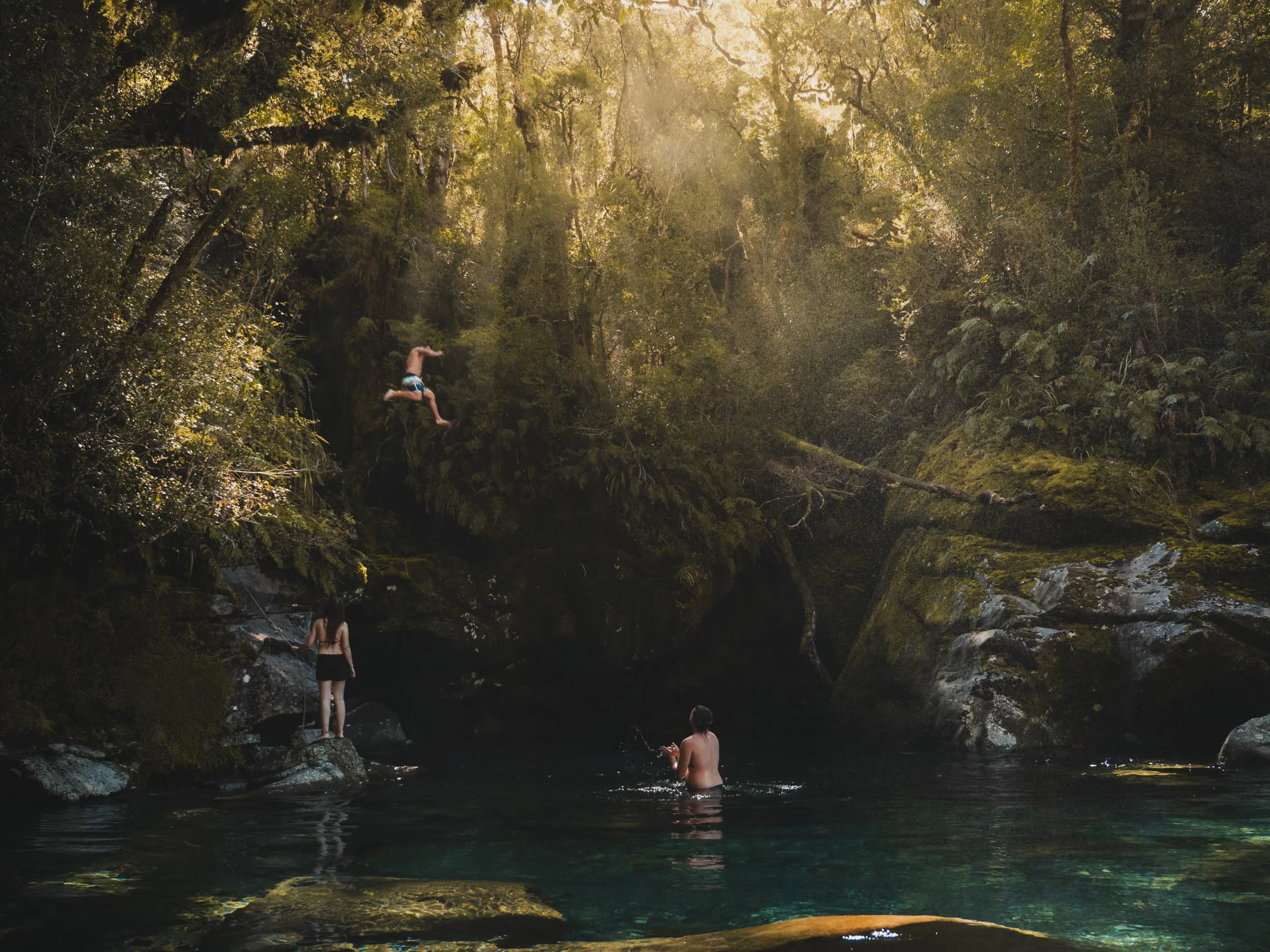 People swimming and relaxing in a river surrounded by lush green trees and sunlight filtering through the branches.