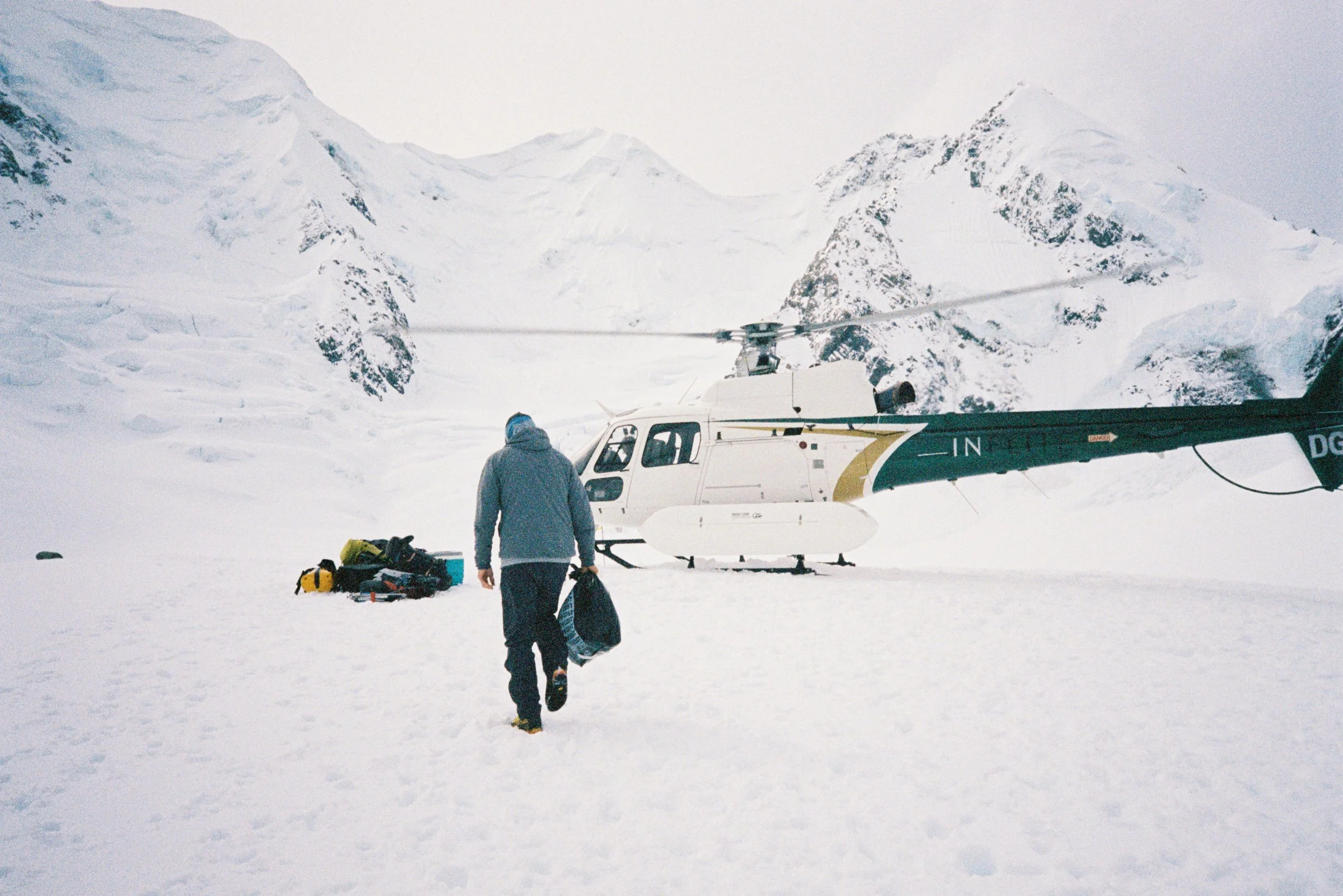 A person in winter clothing carrying gear walks towards a helicopter on snow in a mountainous, snowy landscape with tall, snow-covered peaks in the background.