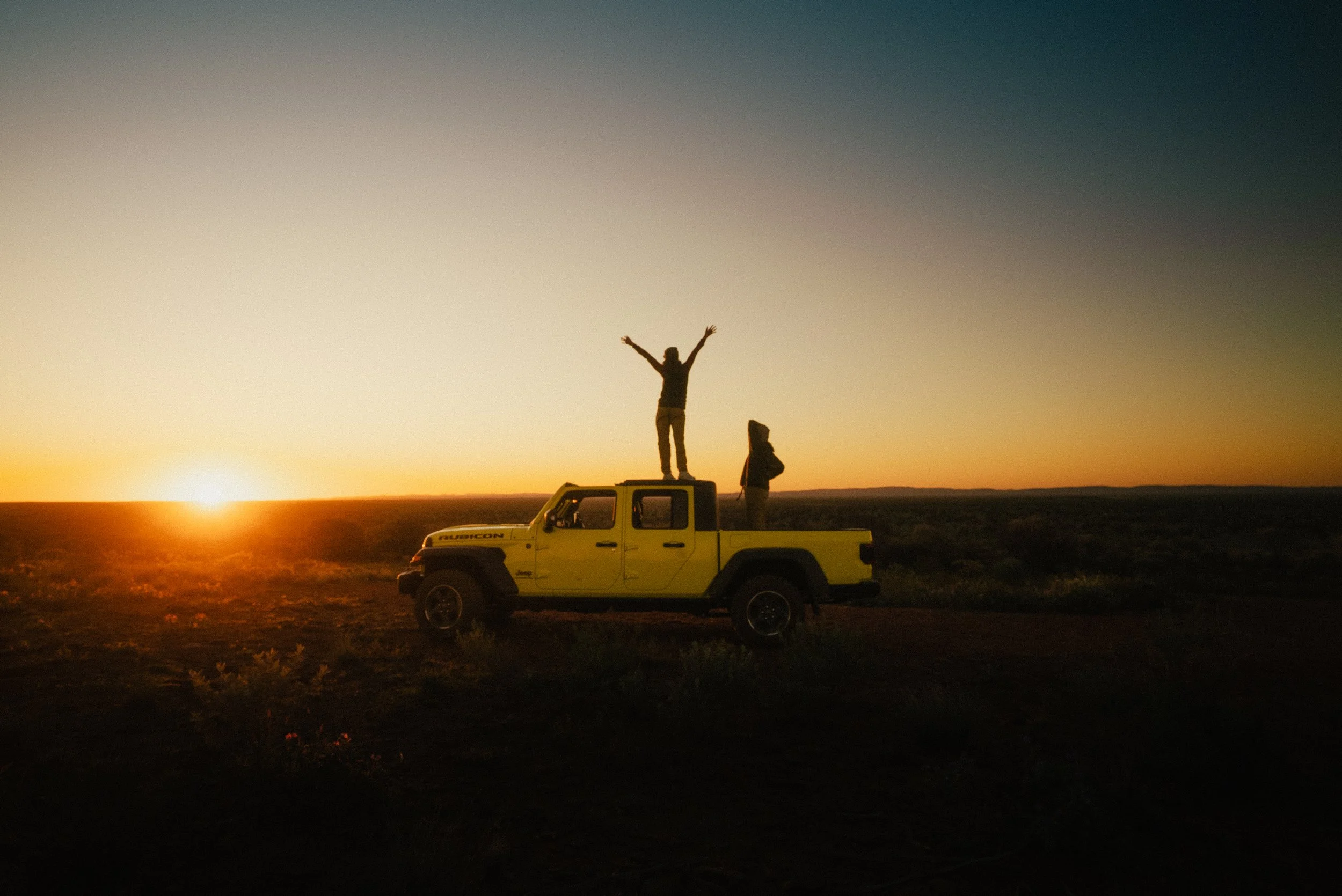 A person standing on the roof of a yellow Jeep Rubicon with arms raised, alongside another person, during a sunset in an open field.