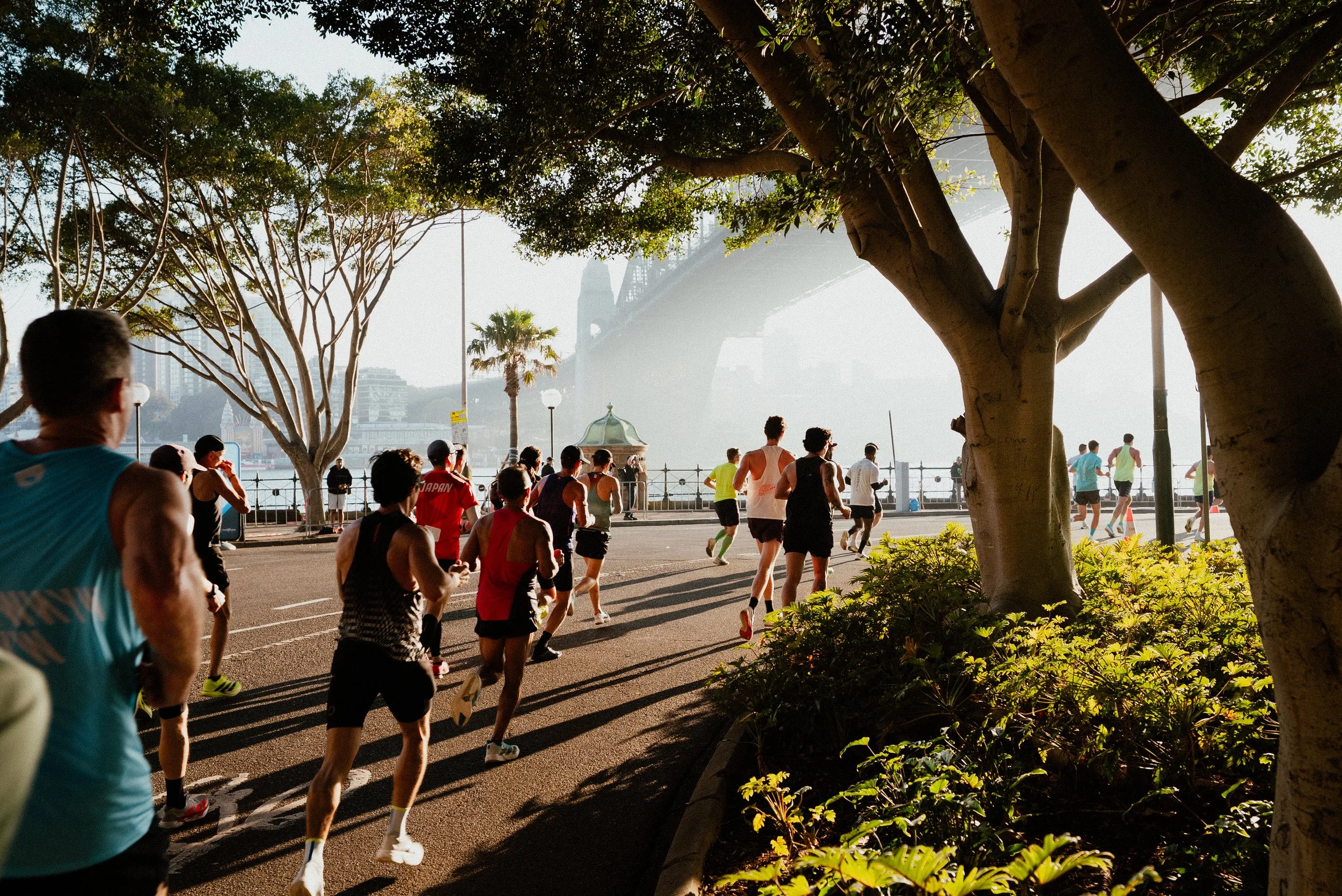 People running along a waterfront park in the morning with trees and city skyline in the background.