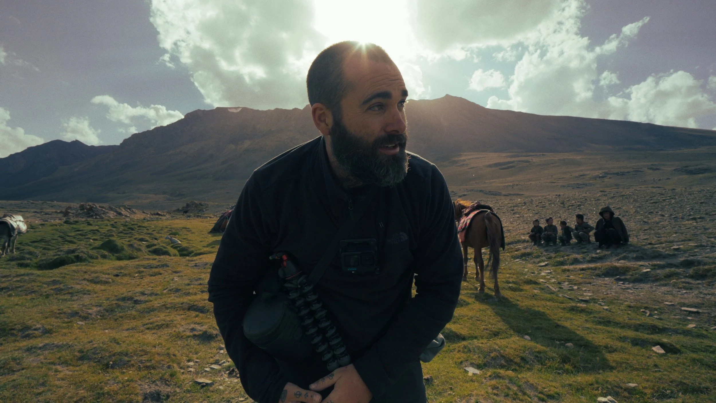 A bearded man wearing outdoor gear, possibly hiking or on an expedition in a mountainous area, with a group of children and horses in the background, under a partly cloudy sky.