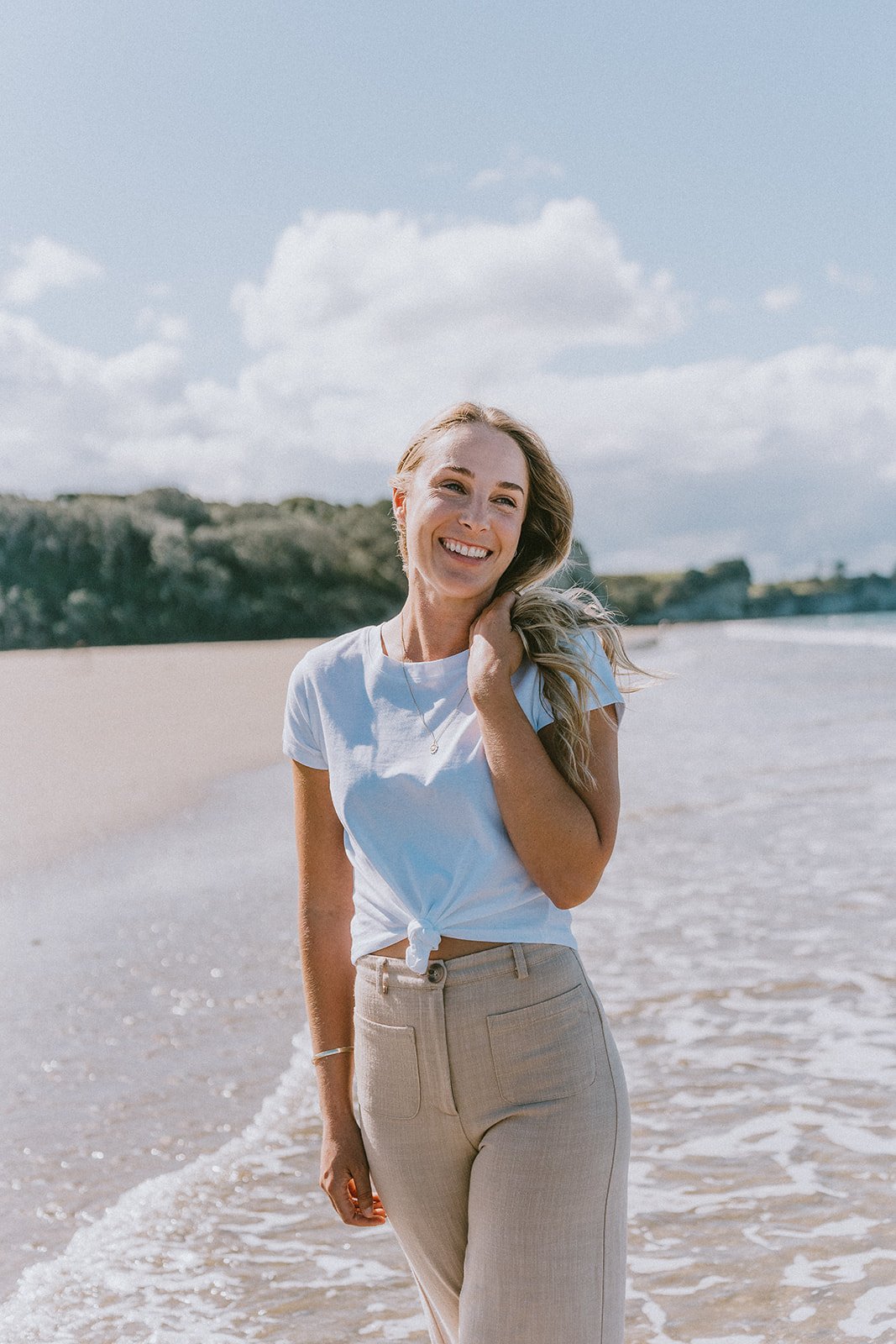 A woman with blonde hair smiling at the camera, standing on the beach near the water with a rocky hill in the background on a sunny day.