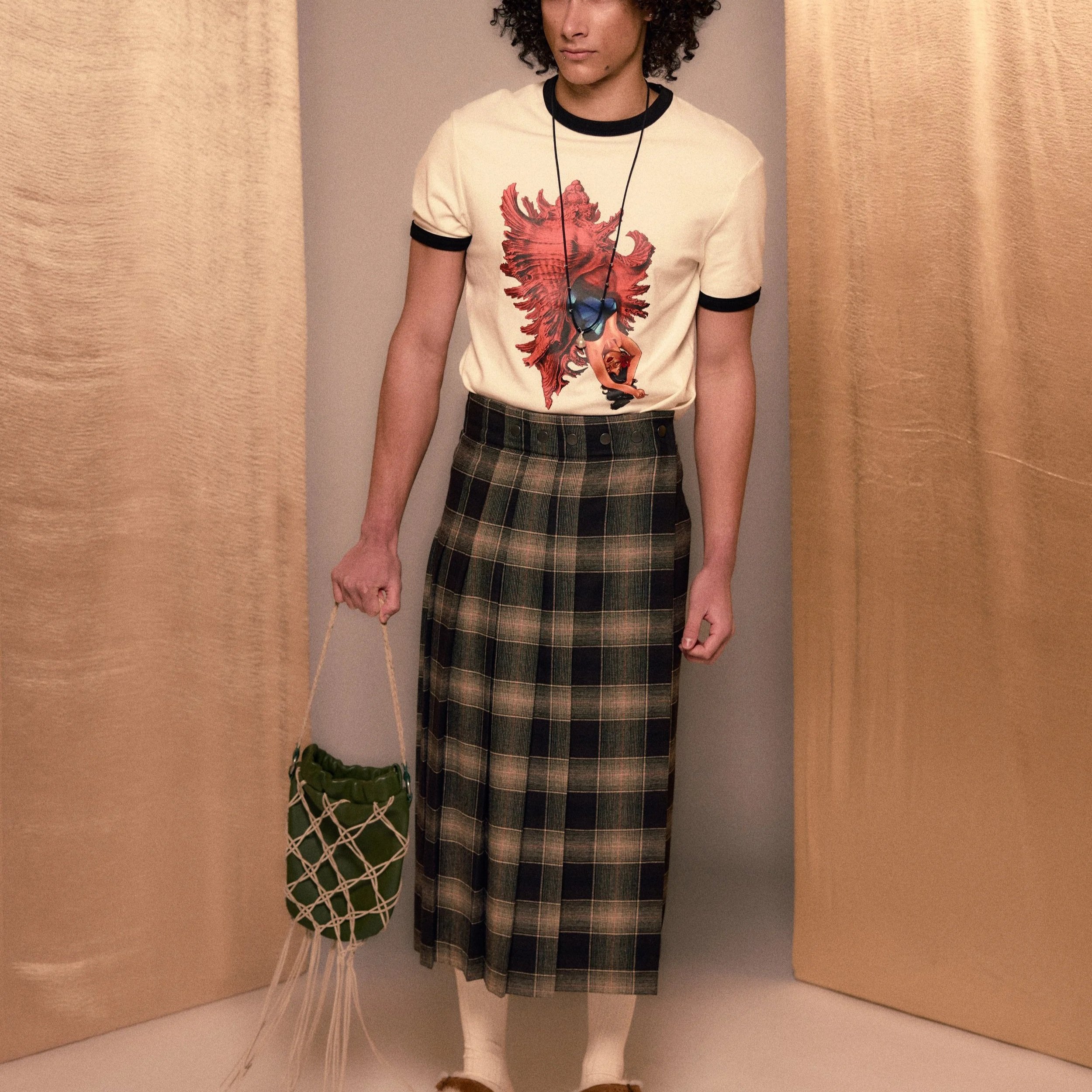 A person with curly hair wears a cream-colored t-shirt with a red, graphic heart and woman design, a plaid skirt, cream-colored tights, and tan shoes. They hold a small green plant in a white, woven netted basket and have a black lanyard around their