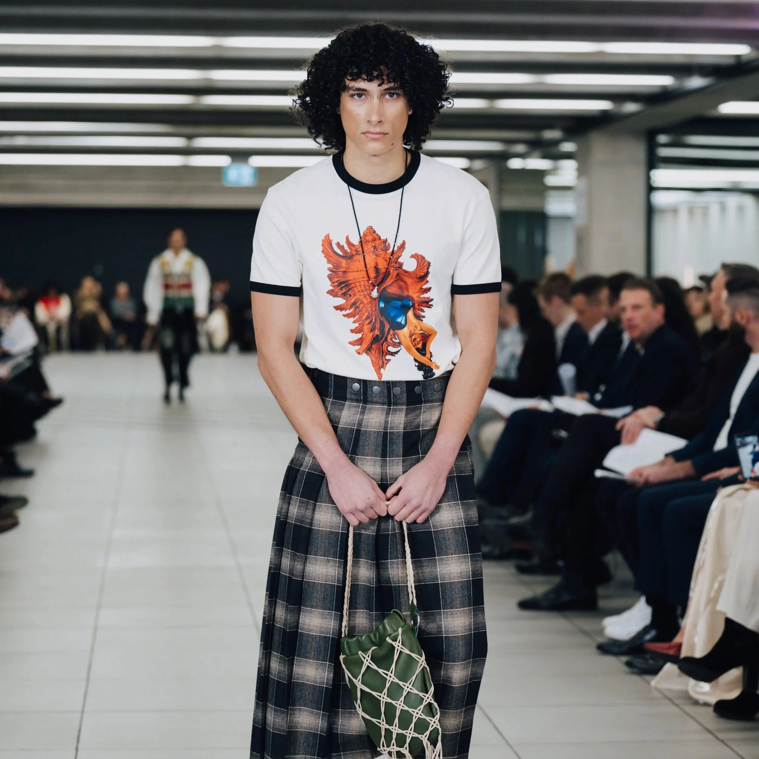 A young person with curly dark hair and light skin standing in an indoor public space, wearing a white t-shirt with a colorful lion graphic, plaid pants, and holding a green bag, with a crowd of people seated in the background.