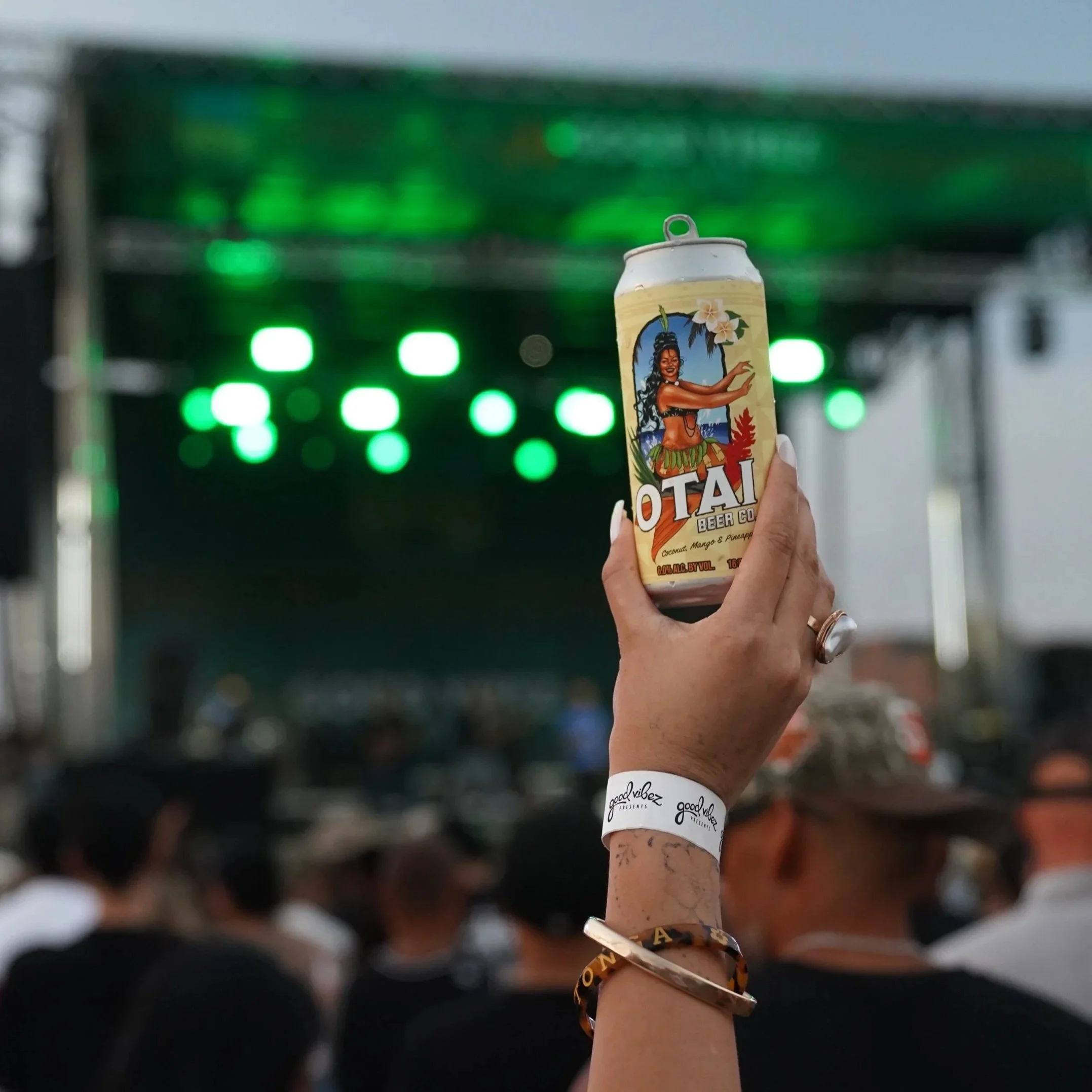 A person holding a can of Hawaii Beer Co.'s Island Style Mango & Pineapple beer in an indoor brewery or bar setting with green lights and blurred background.
