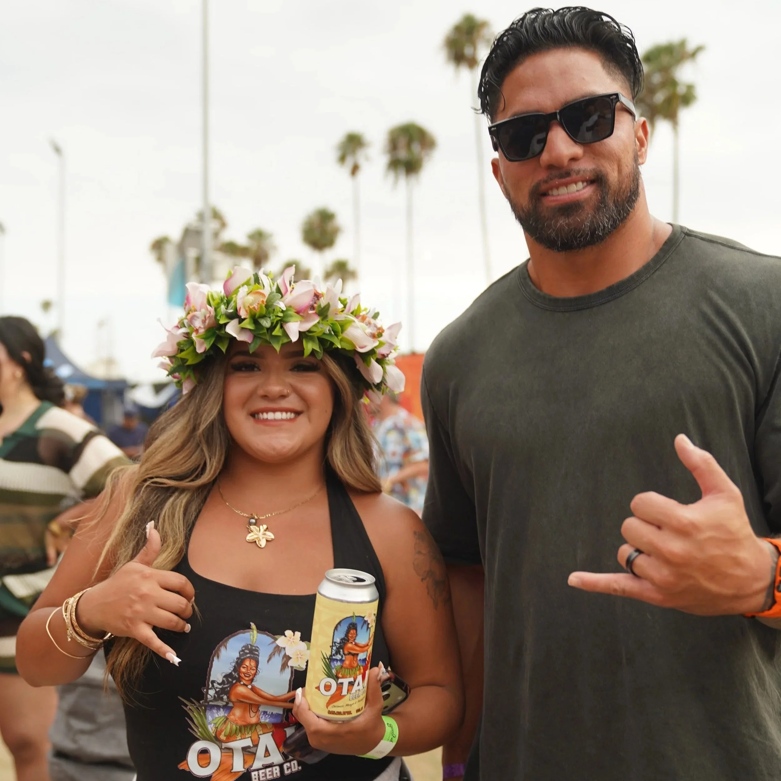 A woman with long, wavy hair wearing a floral crown, black tank top with tropical graphics, and jewelry, standing next to a man in sunglasses and a dark T-shirt, both smiling at an outdoor event with palm trees in the background.