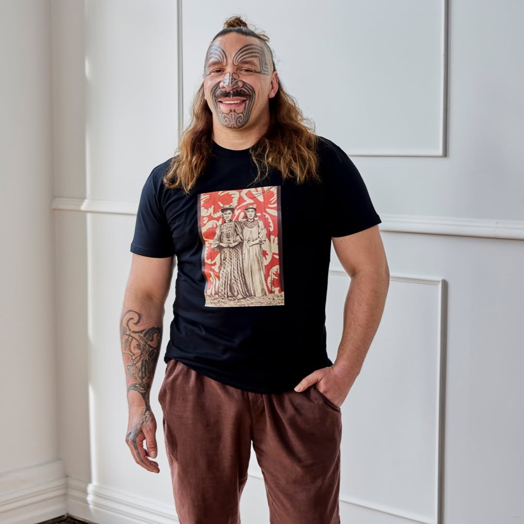 A man with long hair, tattoos, and a floral tattoo on his arm, smiling and standing indoors in front of a white wall, wearing a black t-shirt with a vintage-style photo print of two women and brown pants.