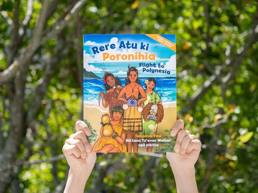 A person holding a colorful children's book titled 'Rere Atu ki Poronihia' with an illustration of four Polynesian children on a beach with the ocean in the background.