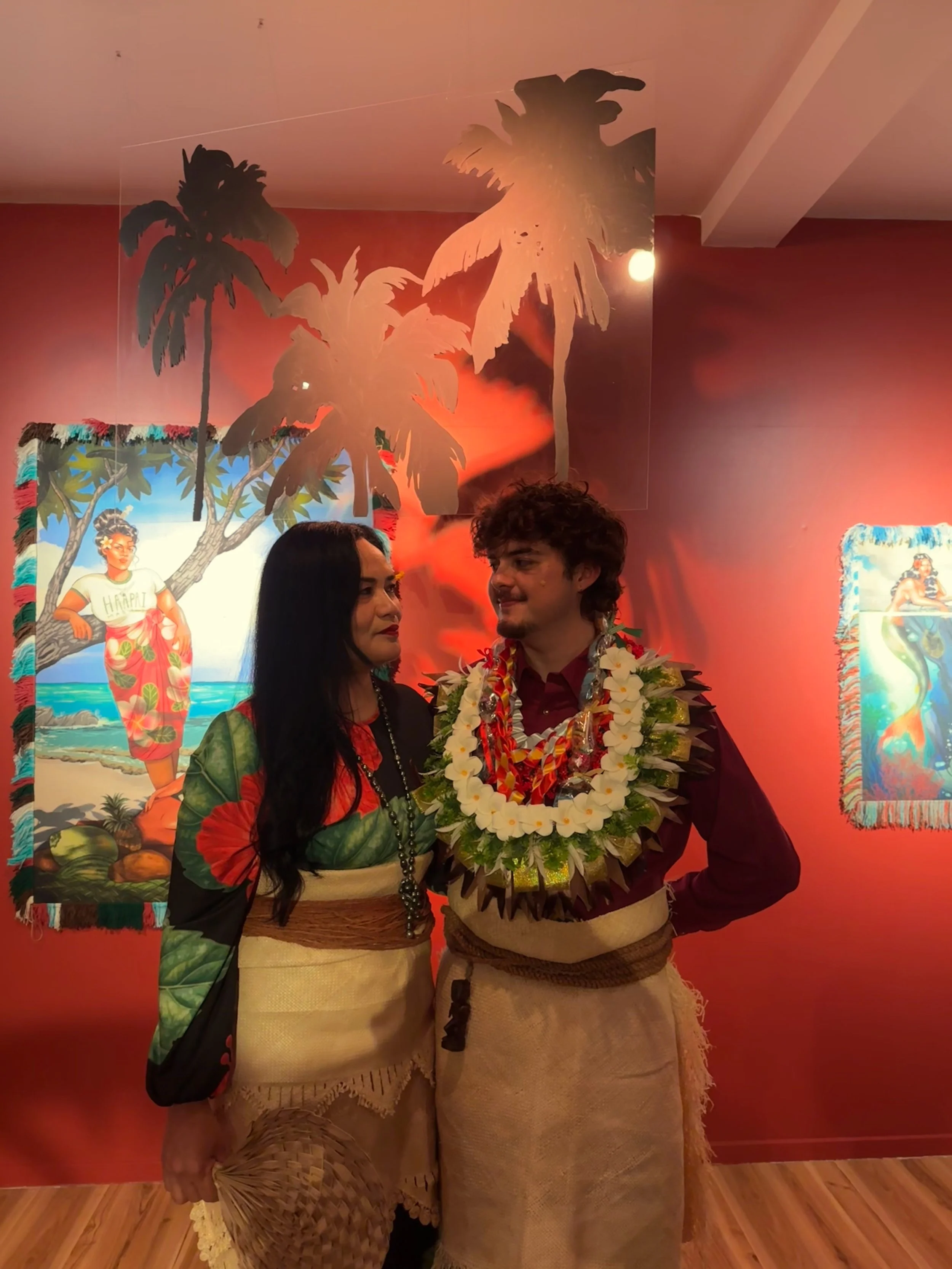 Two people dressed in traditional Polynesian attire at a cultural event, with tropical-themed paintings in the background.