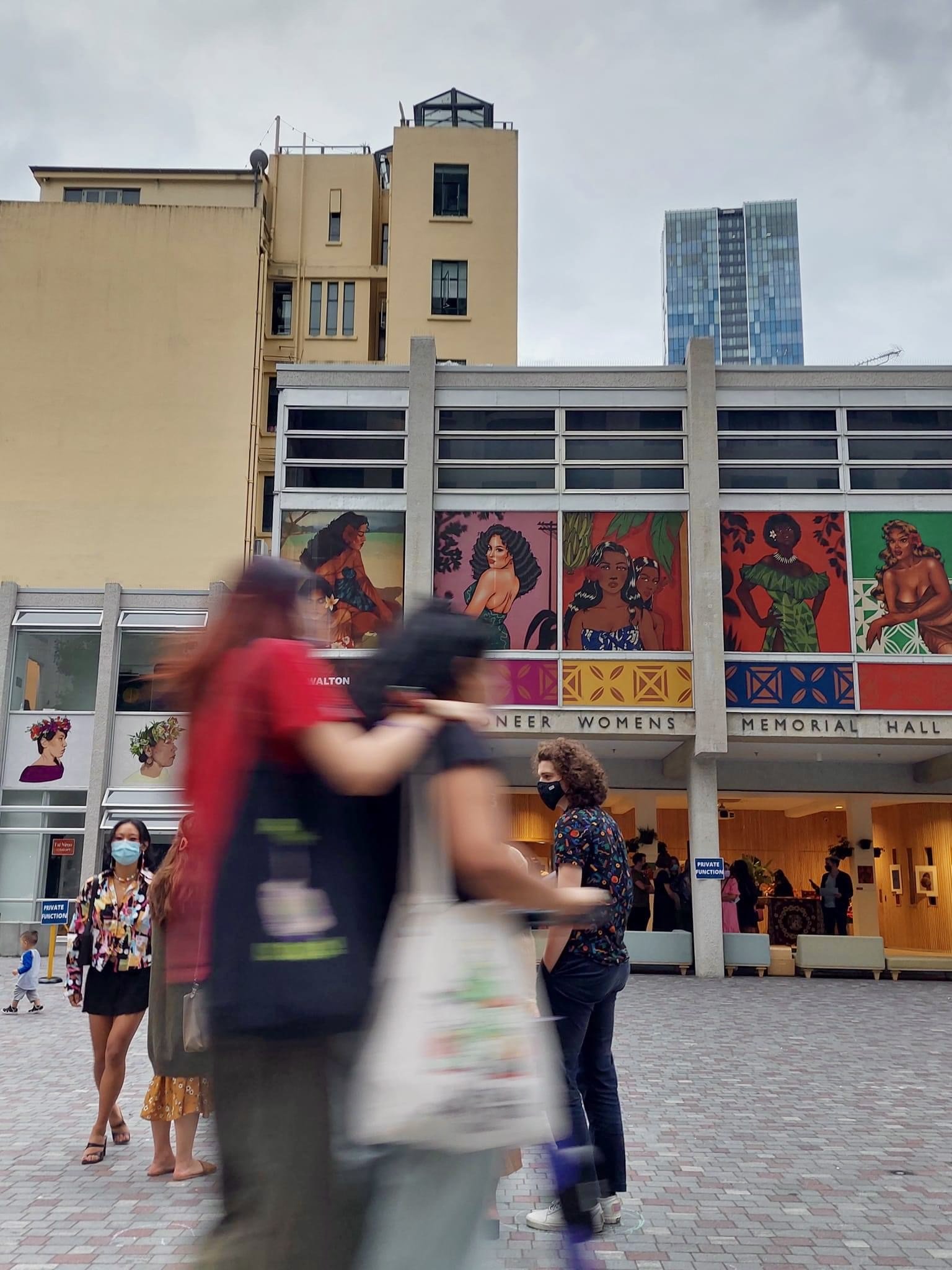 People walking and standing outside a building with colorful murals of women, some wearing masks, under a cloudy sky.