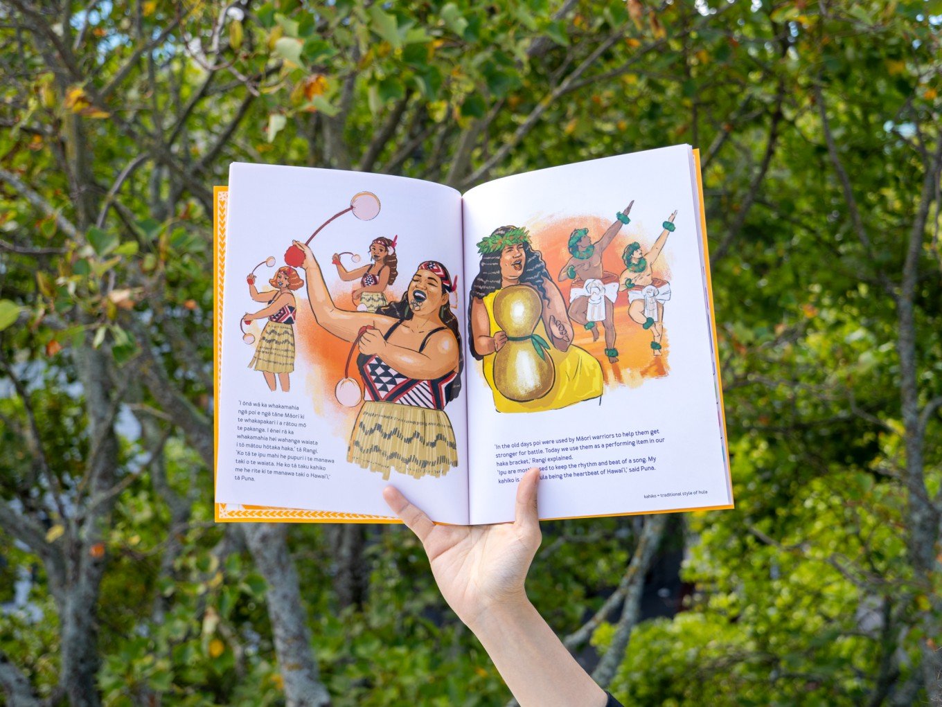 Open book with illustrations of Māori warriors and traditional dance, held outdoors against a background of green trees.