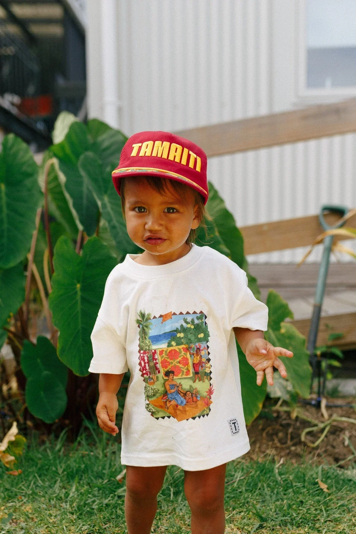 A young child with tan skin and dark hair wearing a red cap with yellow lettering and a white t-shirt with a colorful tropical scene print, standing outdoors in front of green foliage and a wooden bench.