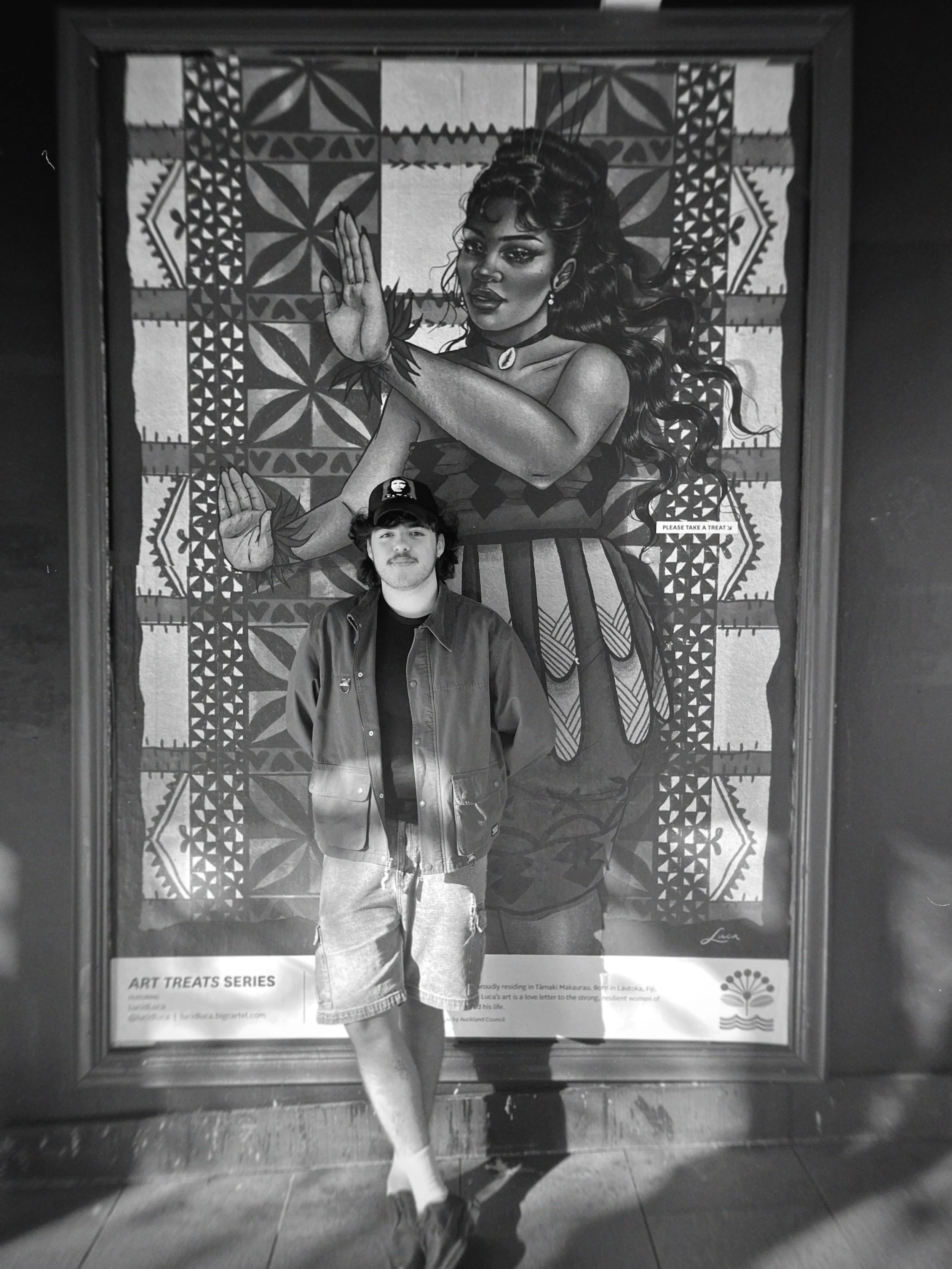 A young man wearing a cap and jacket is standing in front of a large, detailed mural of a woman with long, wavy hair. The woman in the mural is making a gesture with her hand and has a serene expression. The mural has intricate patterns and is part o