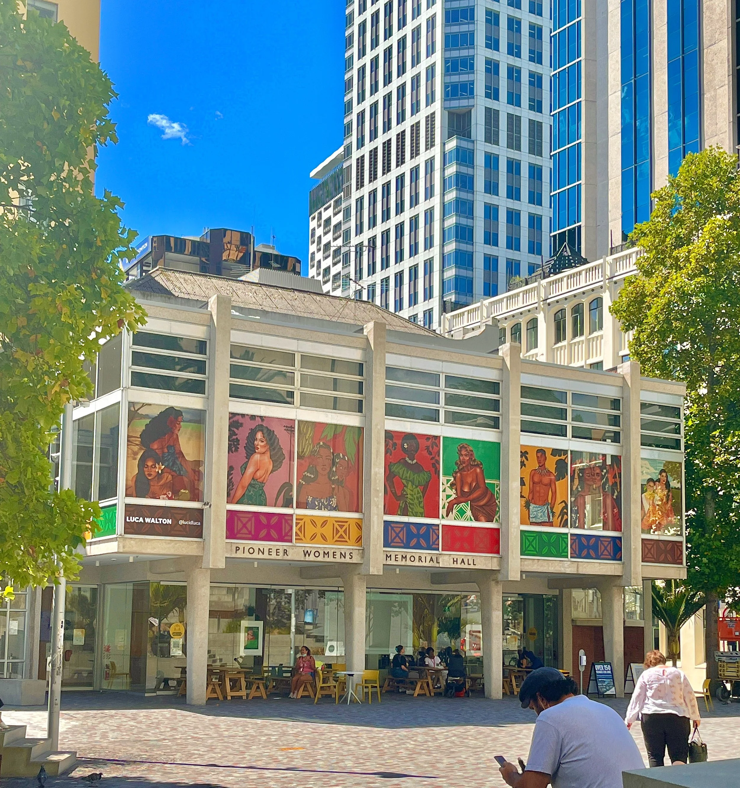 People sitting outside a modern building with portraits of women displayed on the windows, trees on either side, and a clear blue sky overhead.