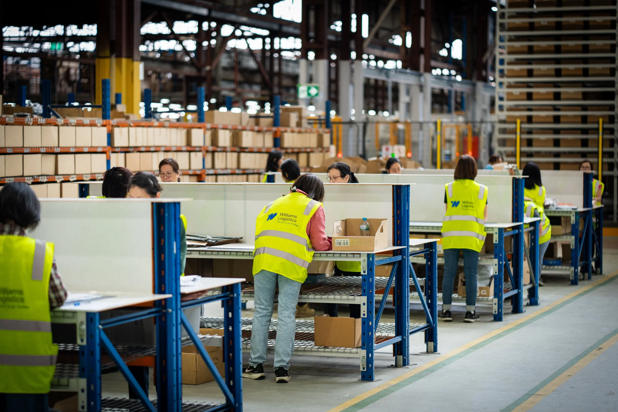 Wide-angle warehouse perspective of 3PL workers focusing on packaing and kitting premium, high end fashion and lifestyle products.