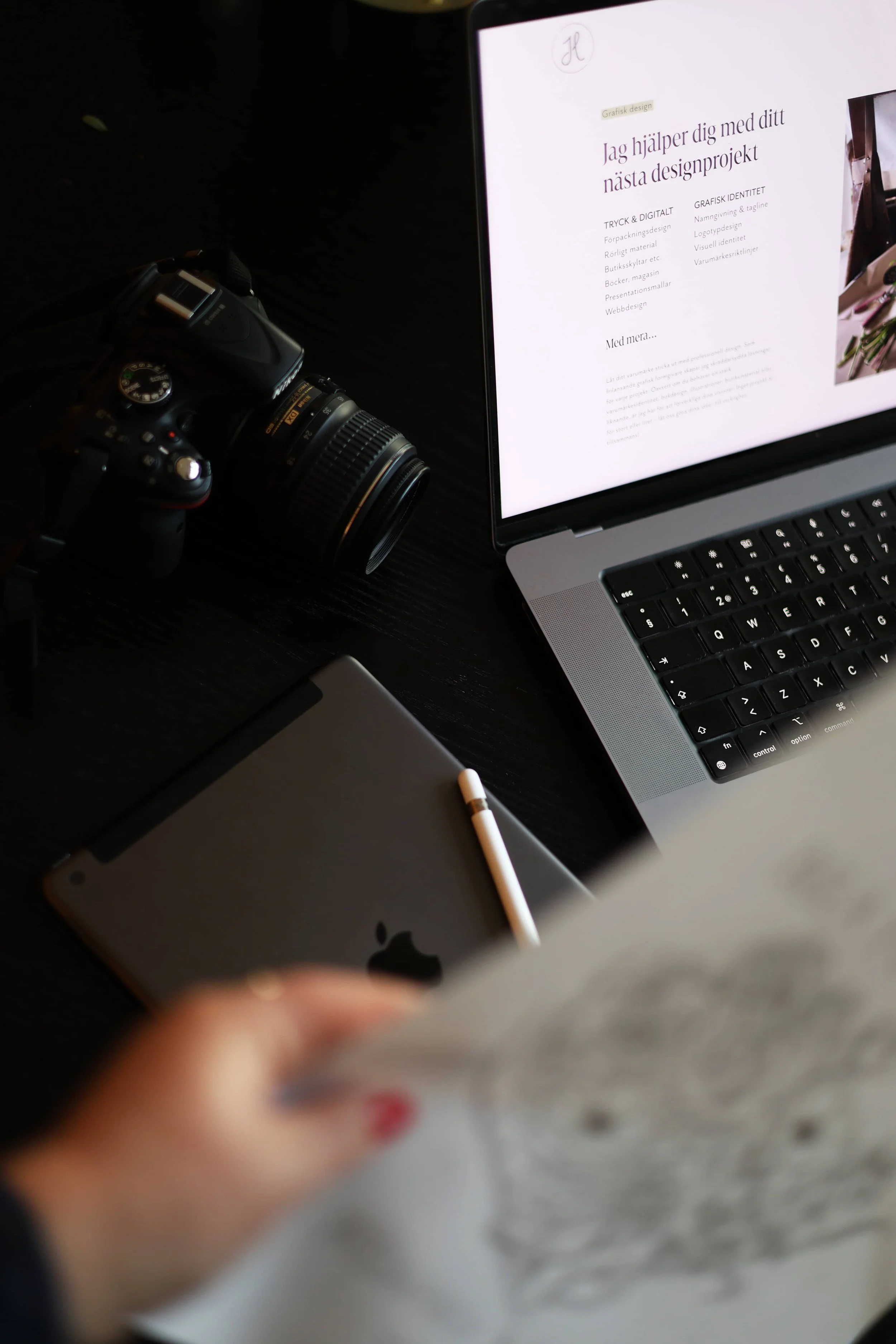 Arbetsbord med kamera, laptop, iPad, stylus och papper. Laptopens skärm visar ett designföretagets webbplats.